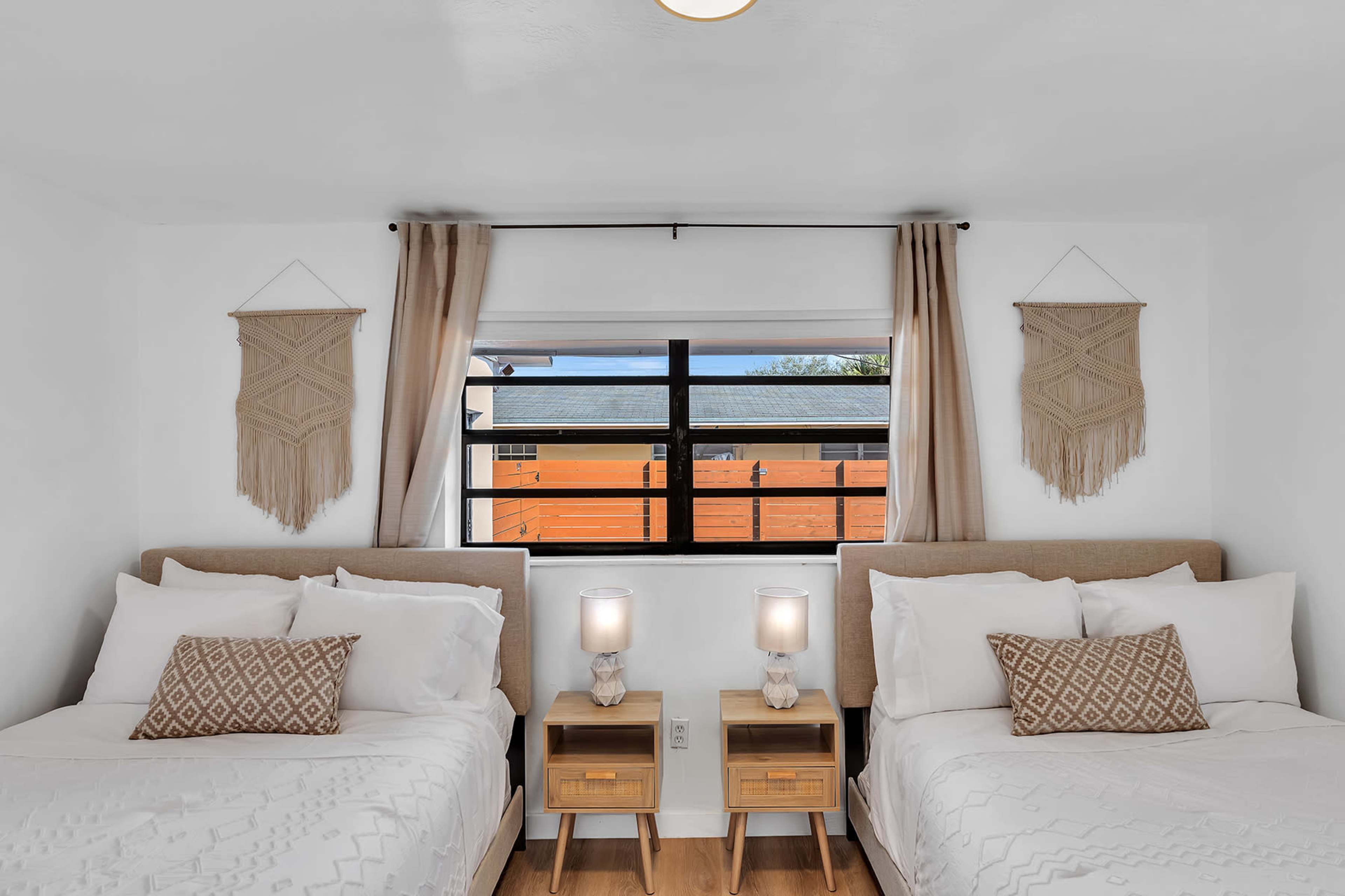 A bedroom with two beds, each featuring white bedding, light wooden nightstands with lamps, and decorative wall hangings above the beds.