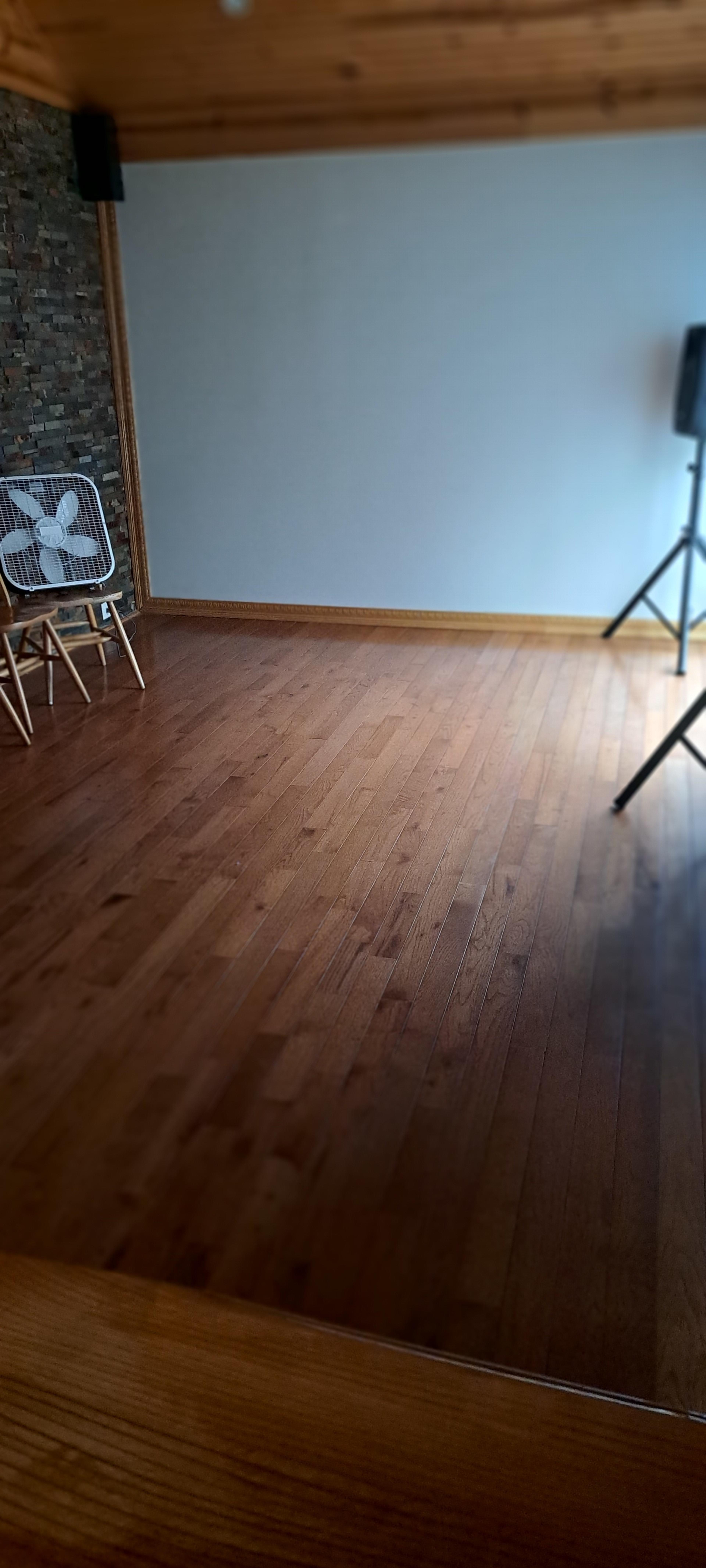 A spacious room with wooden flooring, featuring a fan on a stand and a stone wall section.