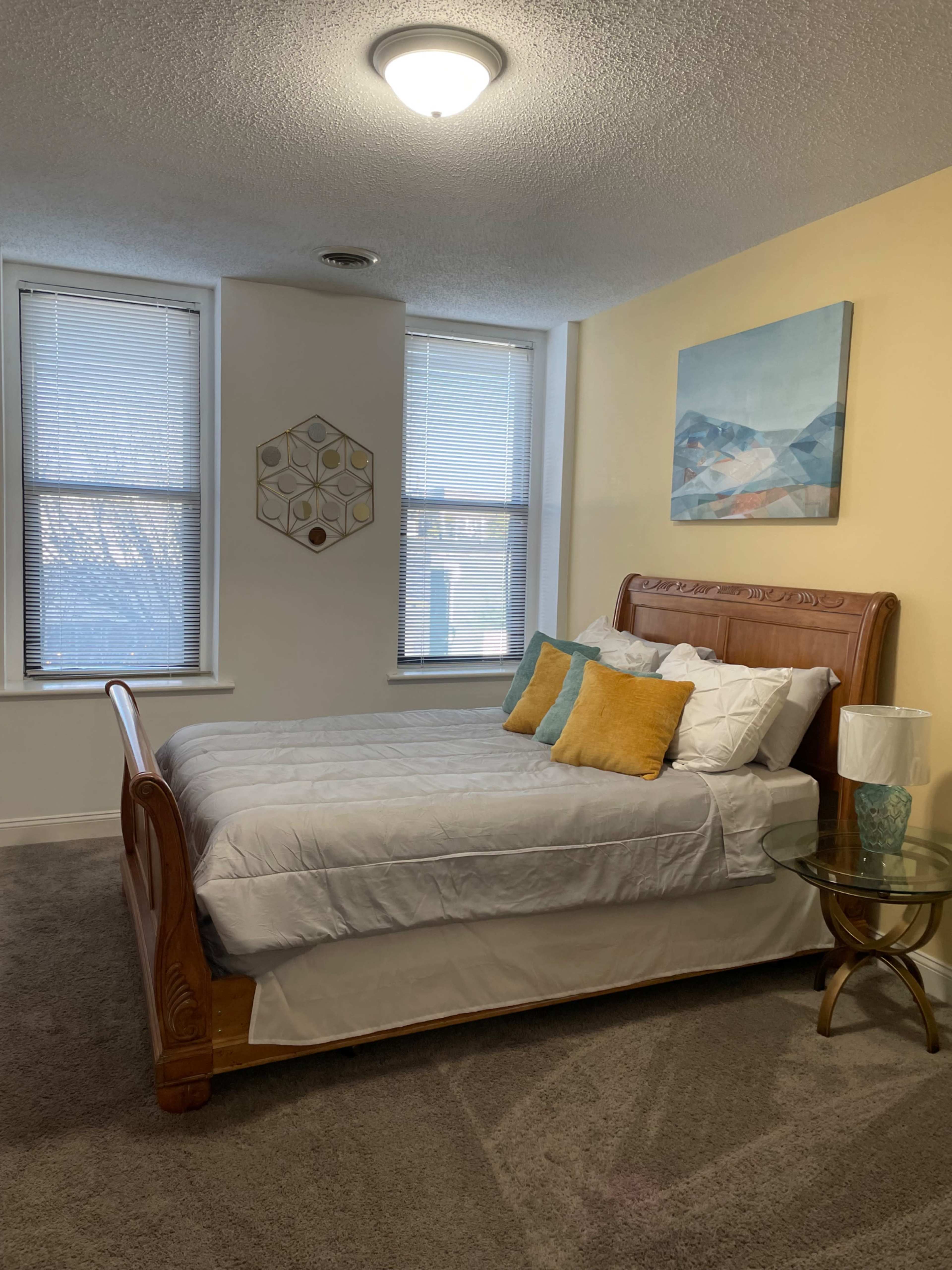 A well-lit bedroom features a wooden bed with gray bedding and colorful pillows, alongside a side table with a decorative lamp.