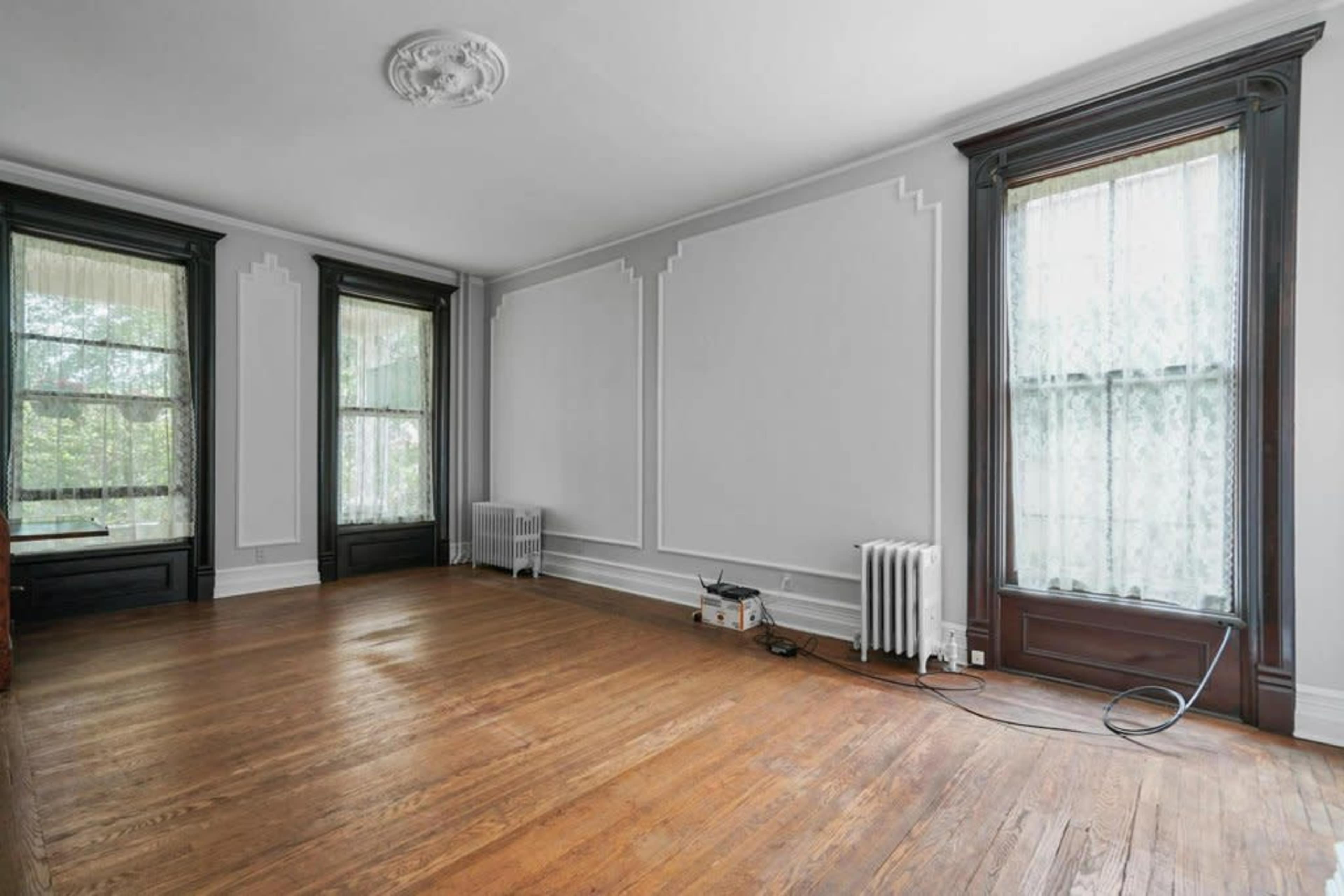 A vacant room with wooden flooring, two windows with lace curtains, and decorative wall molding.