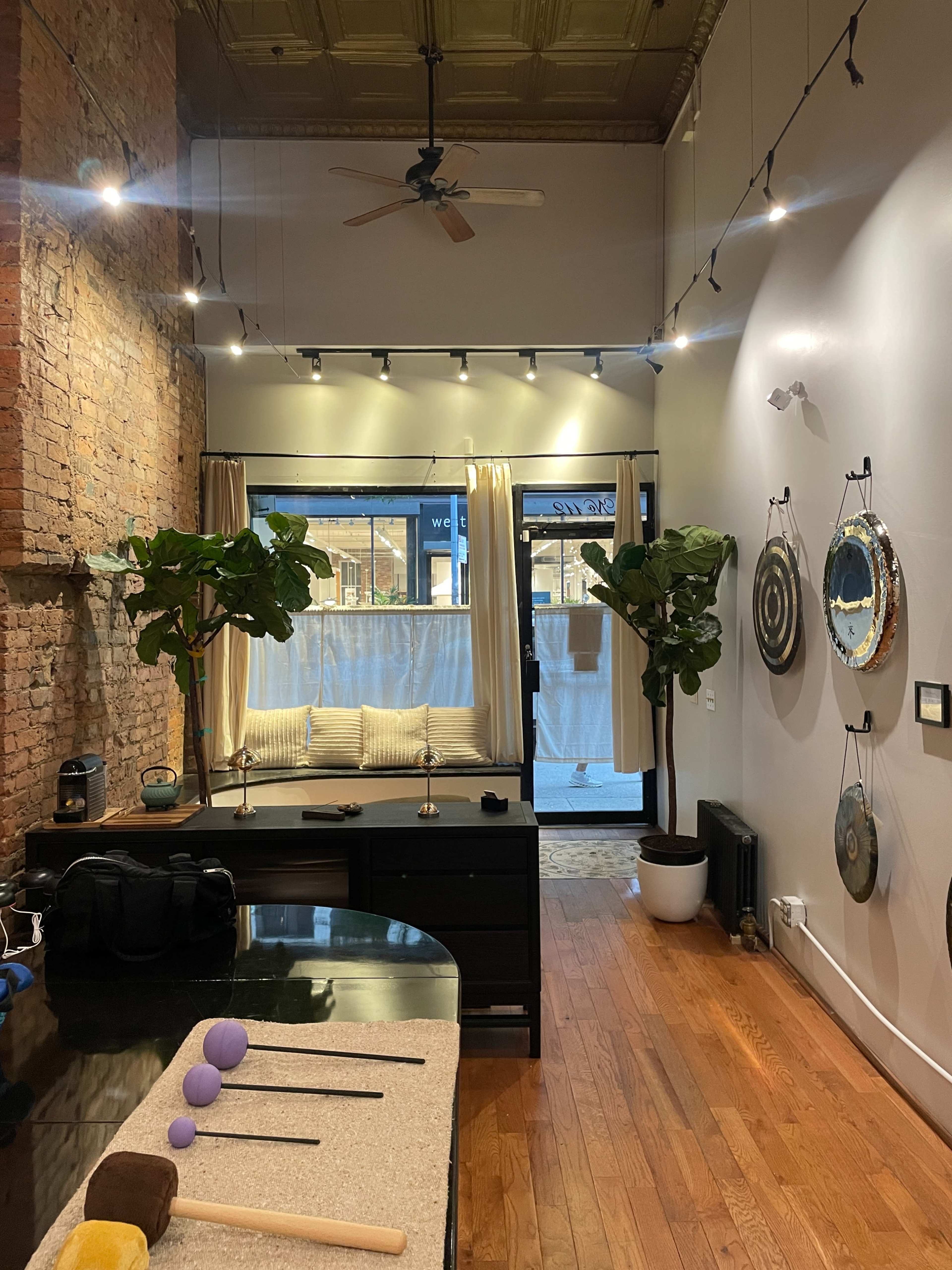 The image shows an interior of a spacious room with exposed brick walls, plants, a seating area by a large window, and several decorative circular objects hanging on the wall.