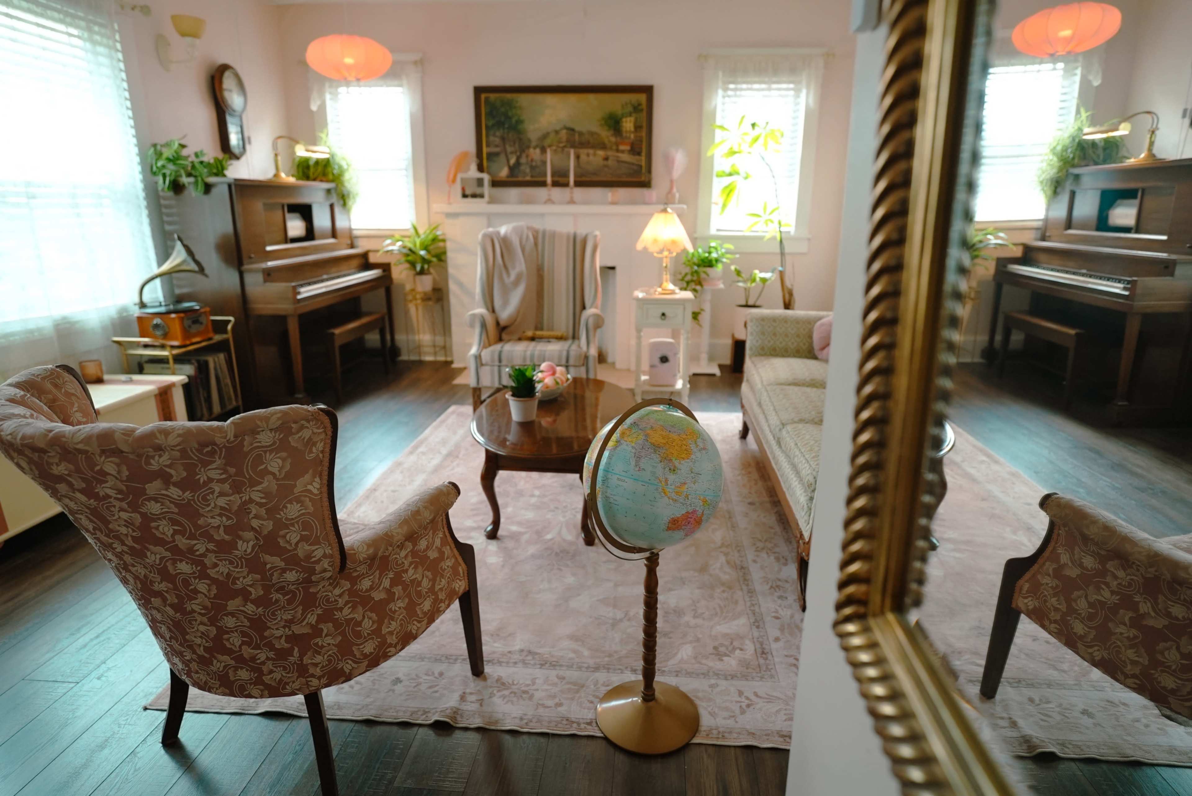 The image shows a cozy living room with two patterned chairs, a wooden coffee table, a globe on a stand, and plants, along with a piano and a painting on the wall.