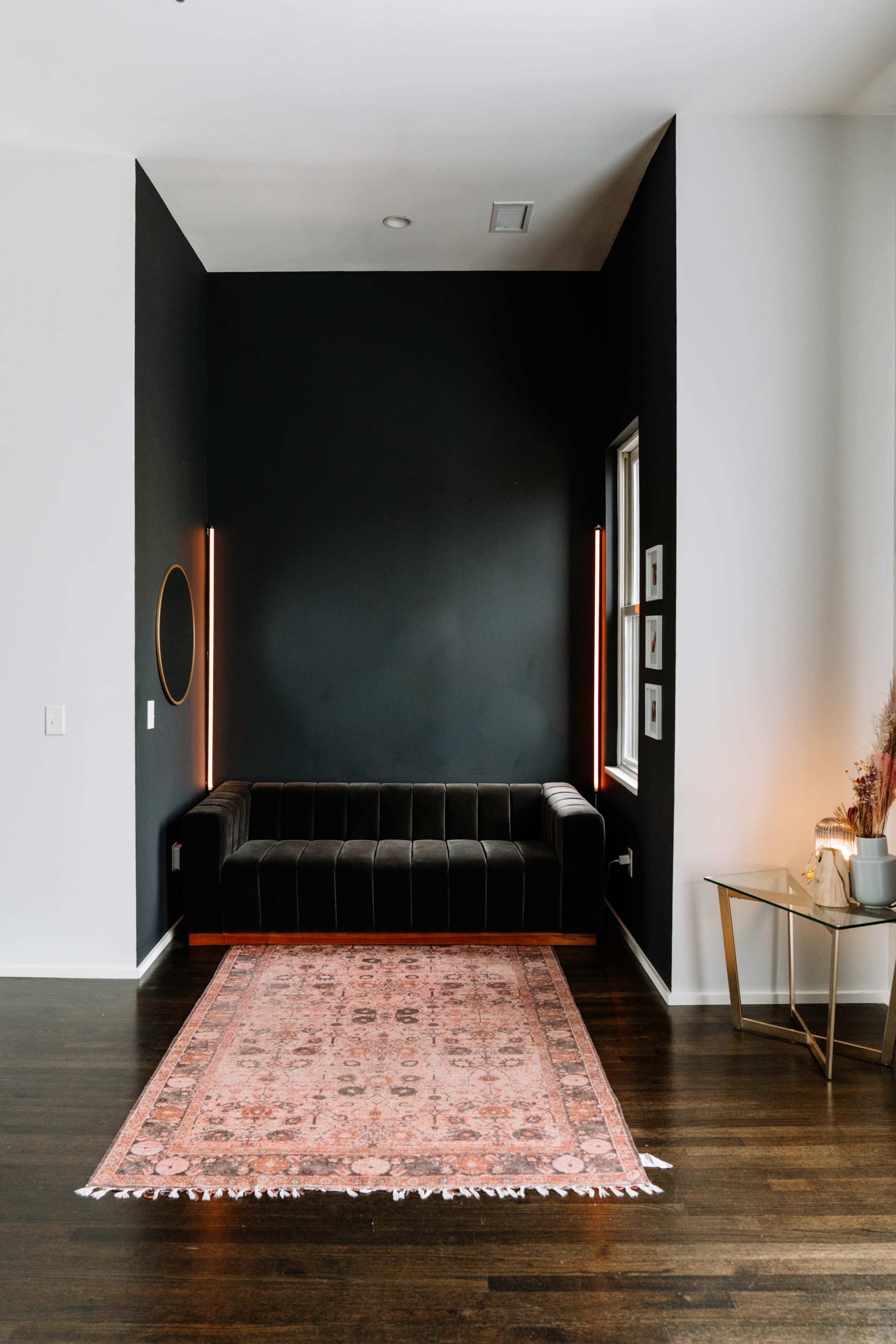 A black sectional sofa is positioned against a dark wall, complemented by a round mirror and illuminated by vertical light fixtures, with a vintage-style rug on the wooden floor.