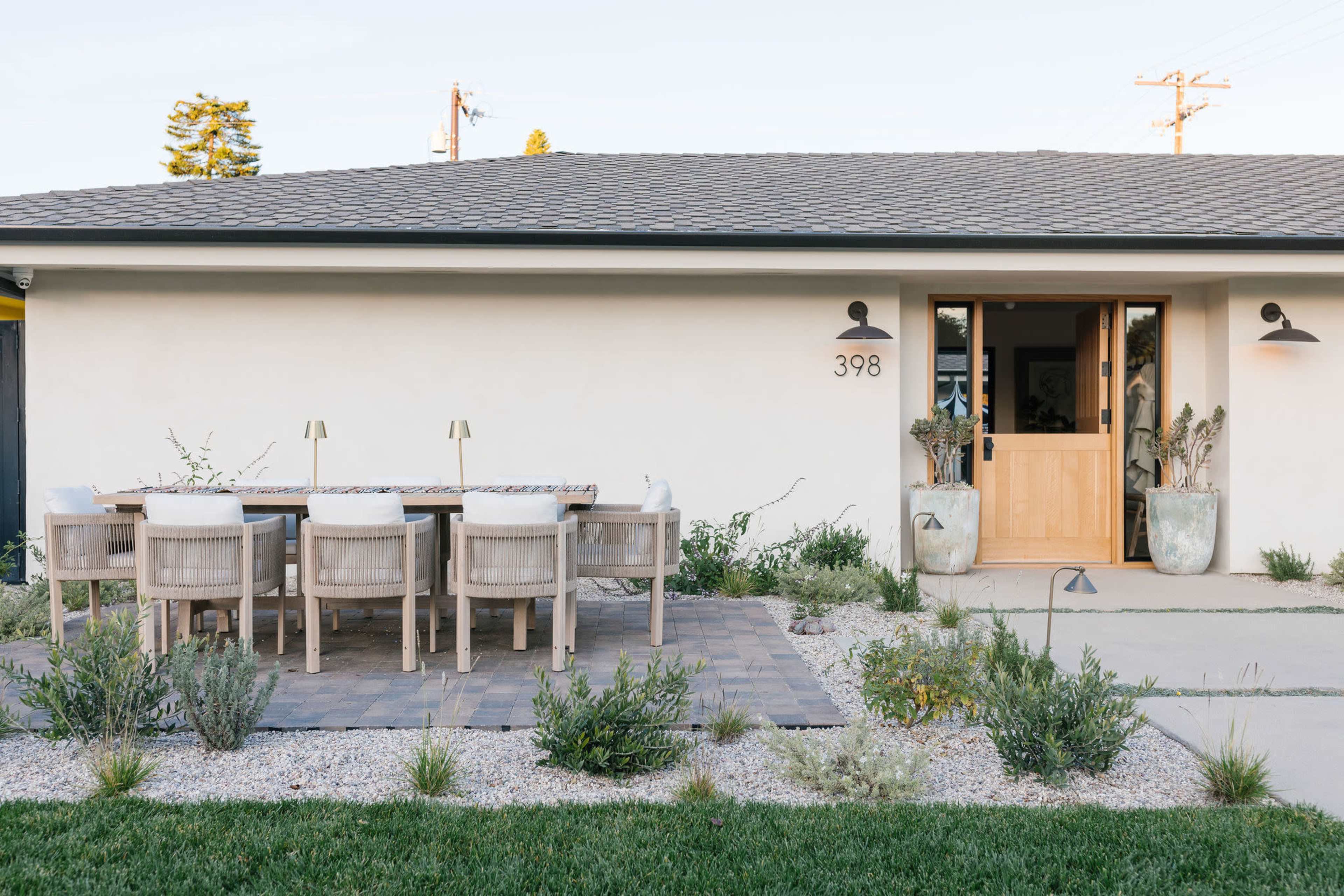 A modern home features a shaded outdoor dining area with a large table and chairs, surrounded by low-growing plants and pebbled landscaping.
