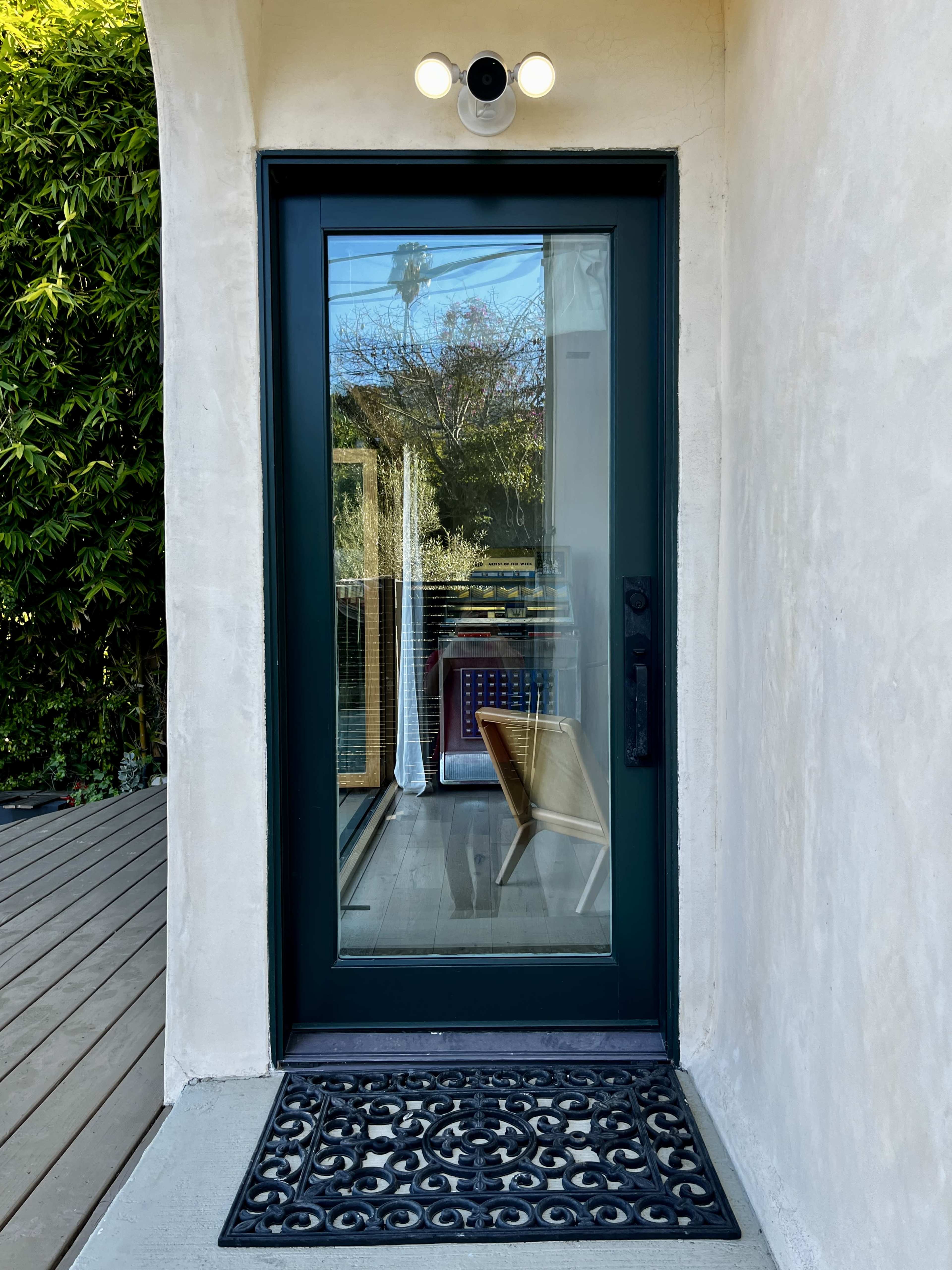 The image shows a dark green front door framed by a light-colored wall, with a decorative doormat and a patio visible through the glass panel.