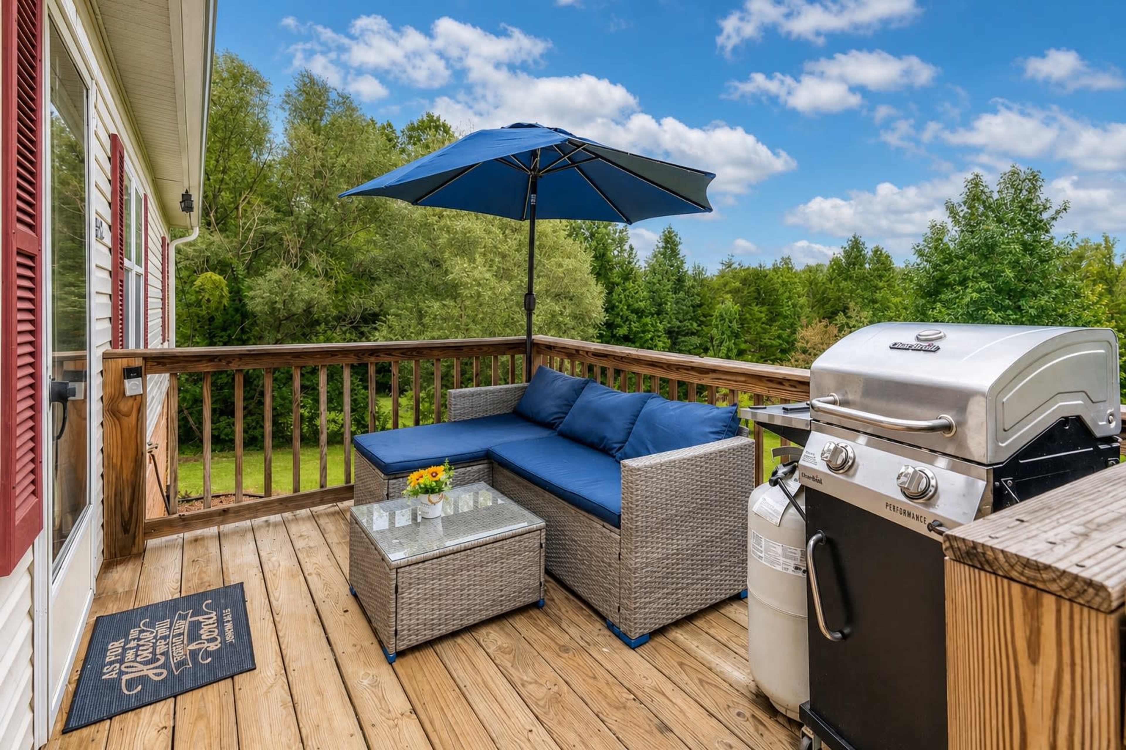 A wooden deck features a couch with blue cushions, a glass-topped table with flowers, a grill, and an umbrella, overlooking a lush green landscape.