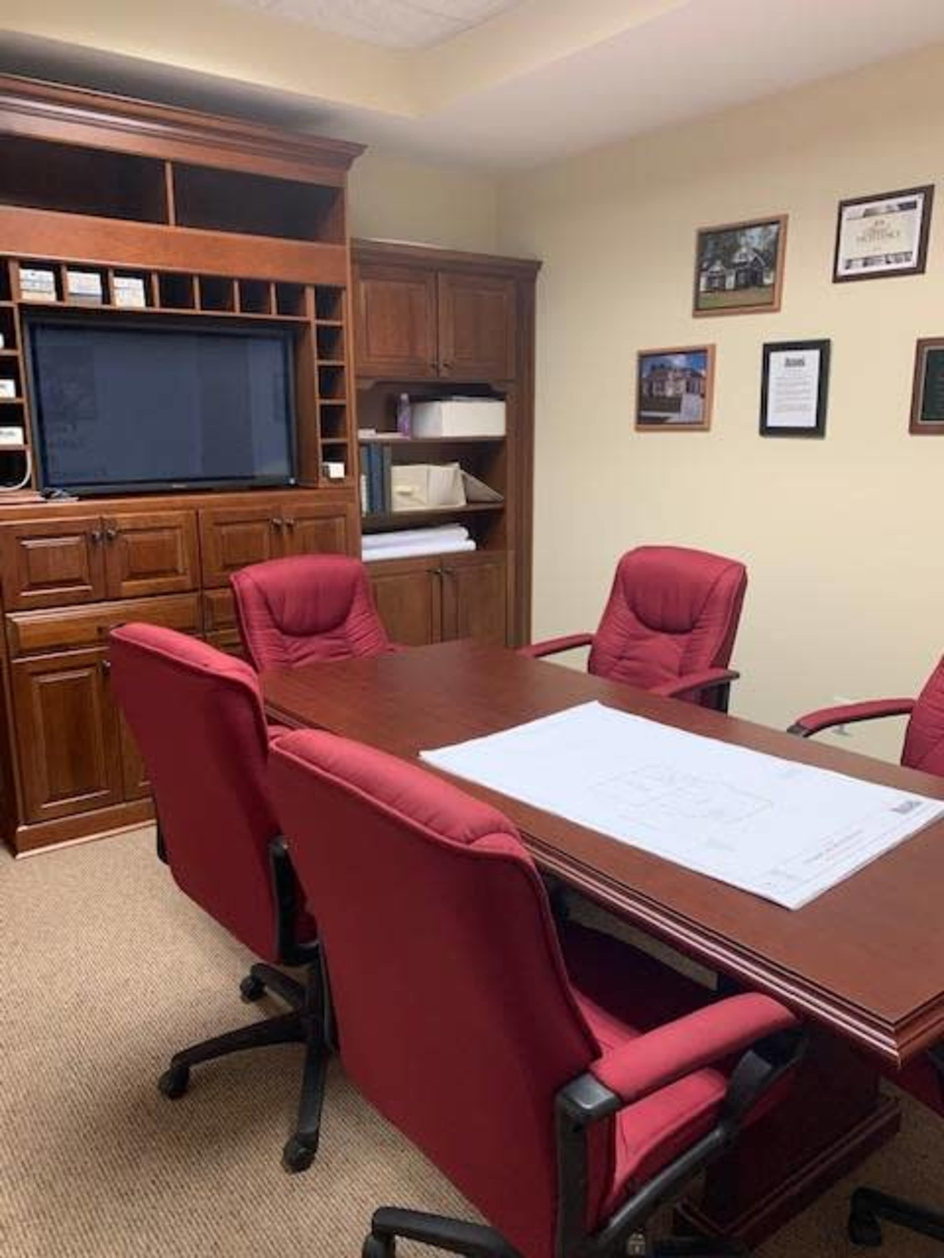 A conference room features a rectangular table surrounded by five red chairs, with a wooden cabinet and wall-mounted TV in the background.