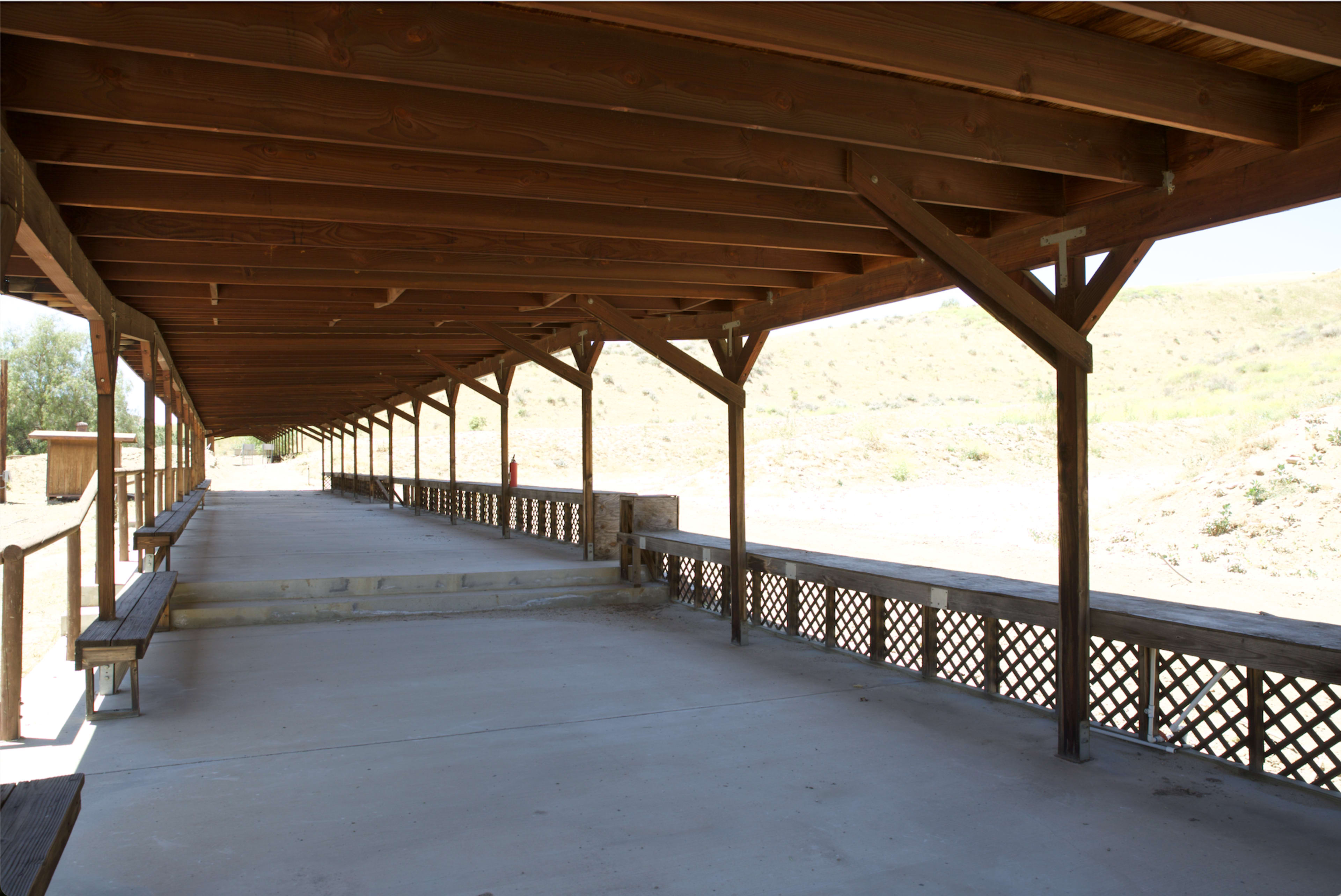 The image shows an empty wooden shelter with a sloped roof, featuring a concrete floor and benches along the sides.