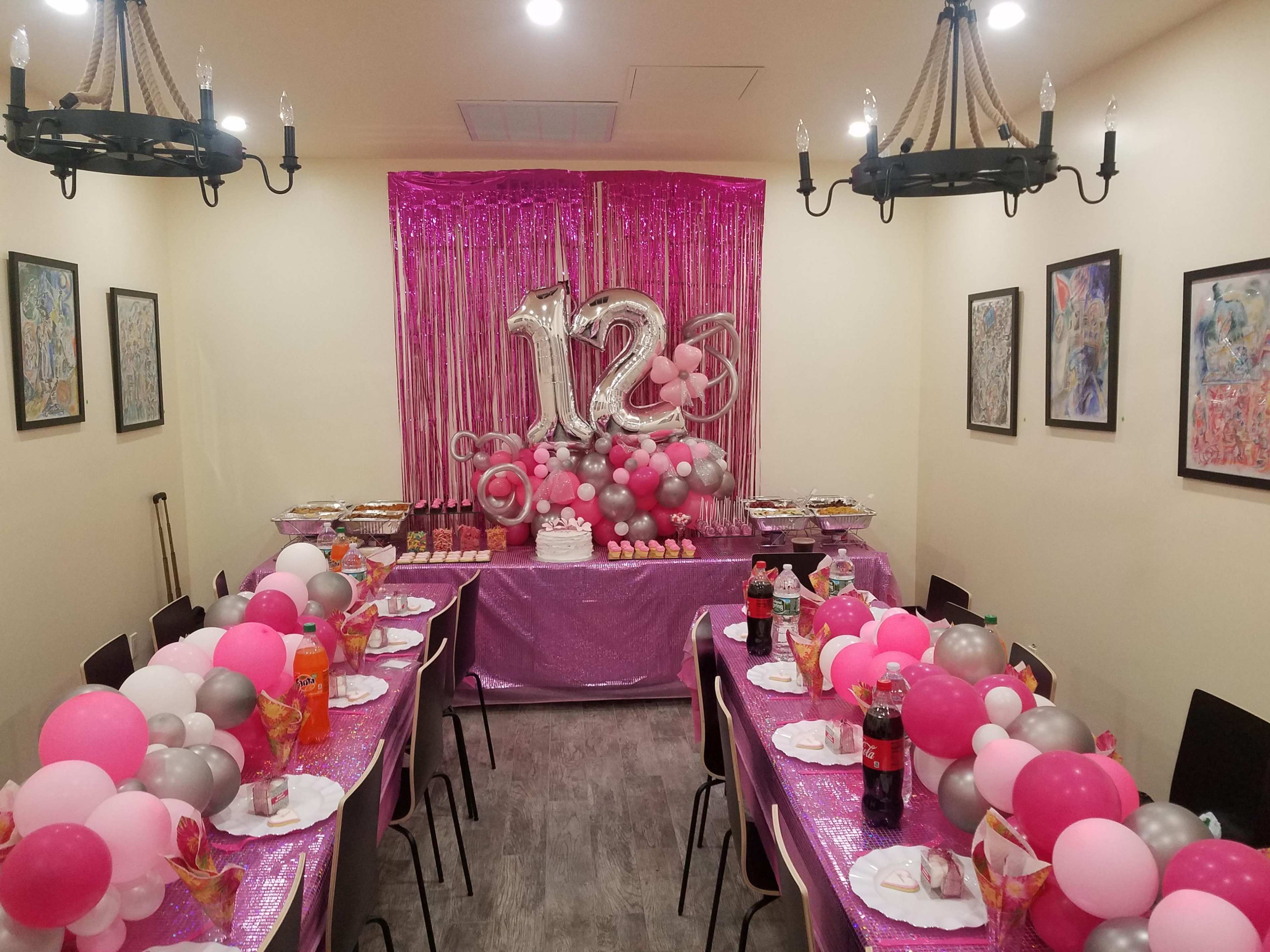 A decorated dining area features a long table set for a celebration, adorned with pink, gray, and silver balloons, a cake, and various food platters.