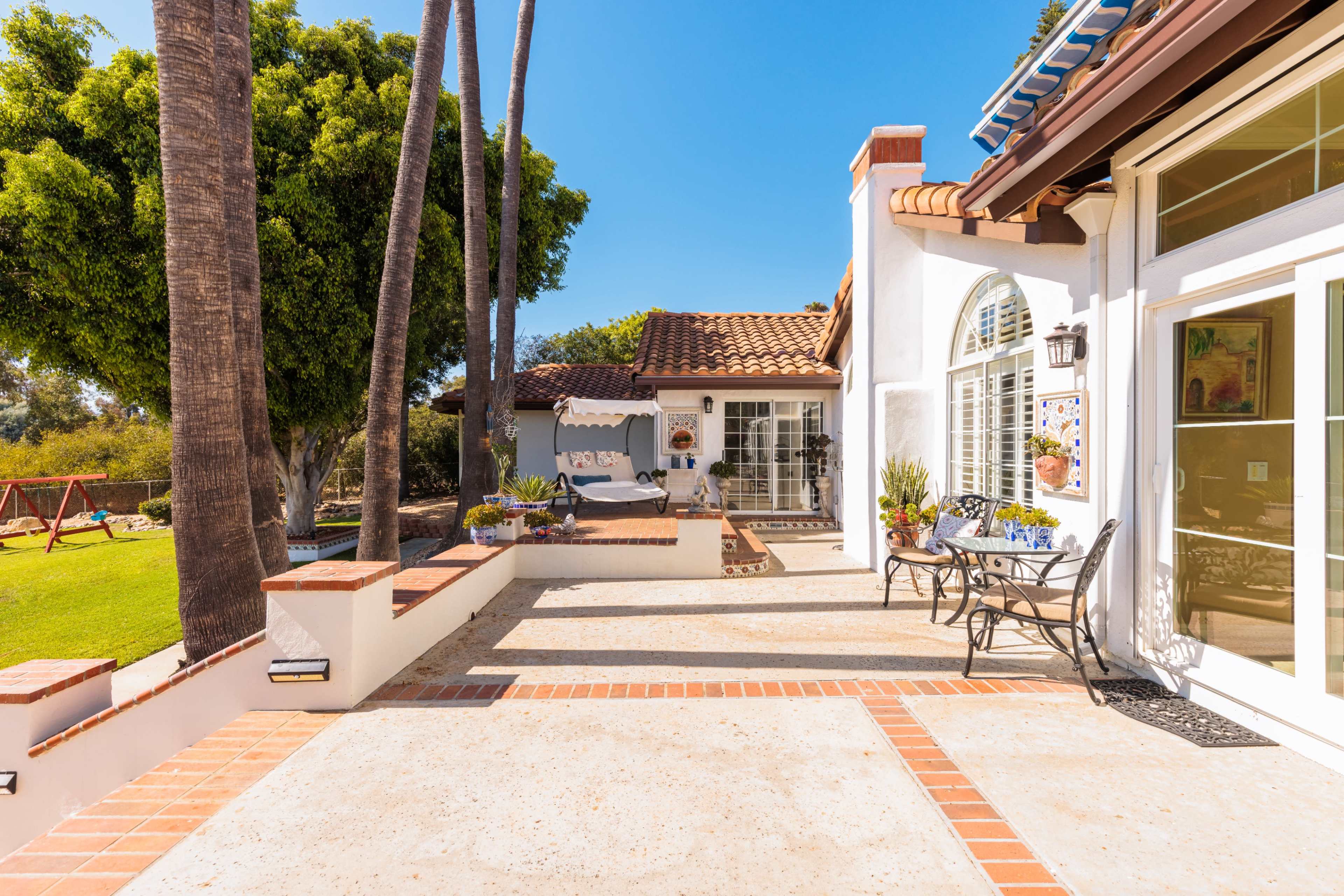 A sunny courtyard area with stone flooring, bordered by tall palm trees and a view of a swing set in the background.