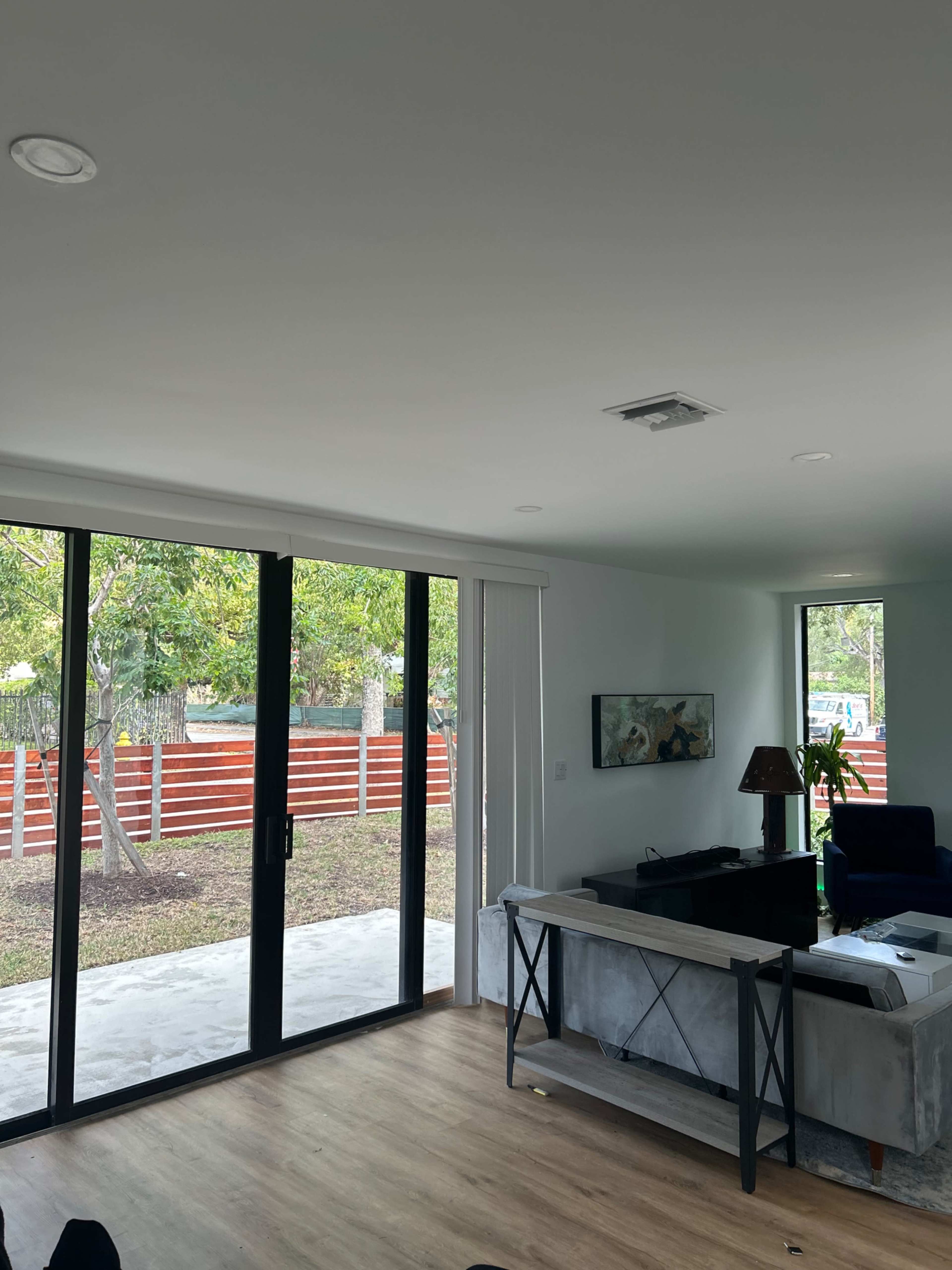 The image shows a minimalist living room with large glass doors opening to a view of a backyard fenced with wood.