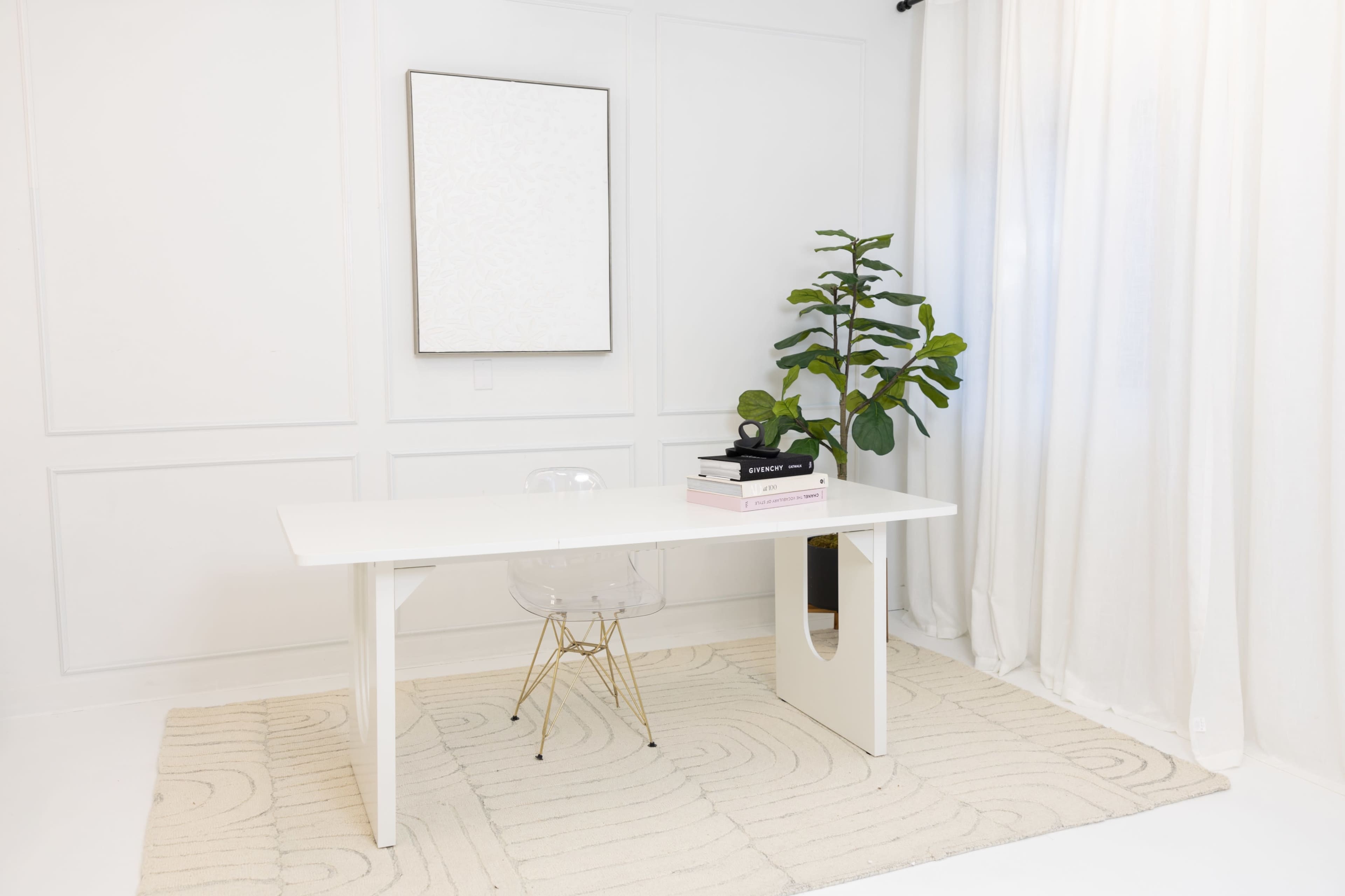 A minimalistic office space features a white desk, a clear chair, a potted plant, and a large window with sheer curtains.