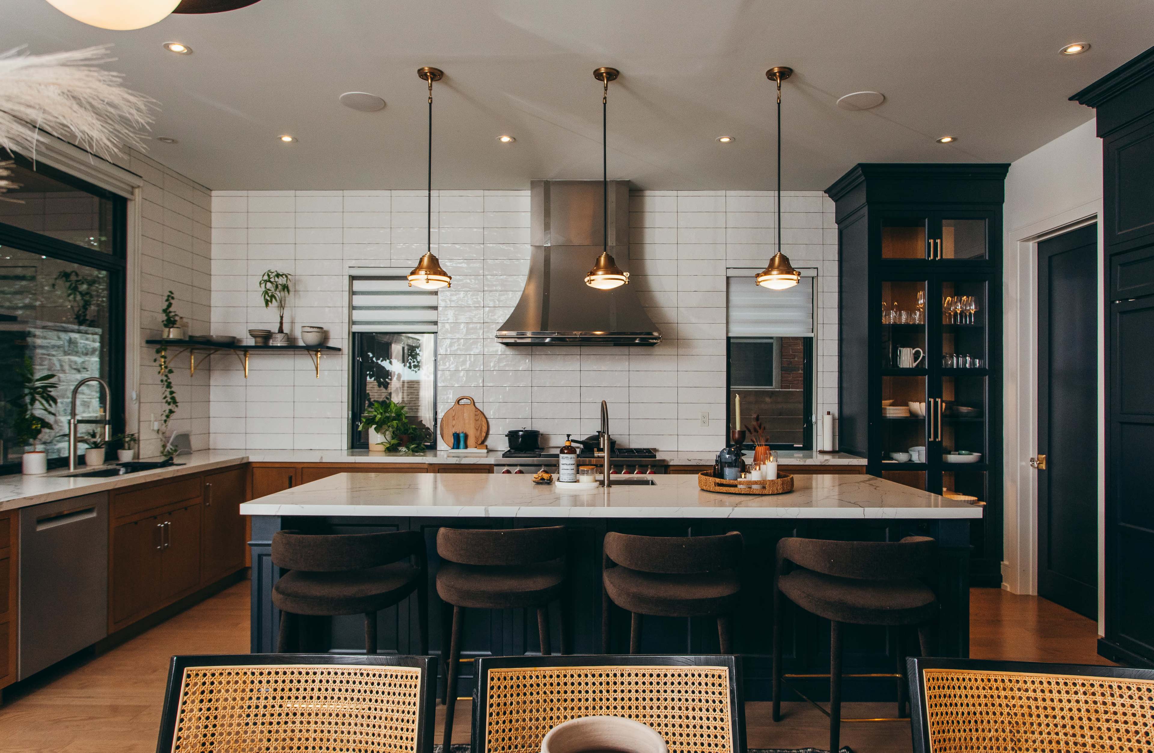 The image shows a modern kitchen with a central island, bar stools, a stainless steel range hood, and a combination of dark cabinetry and white tile backsplash.