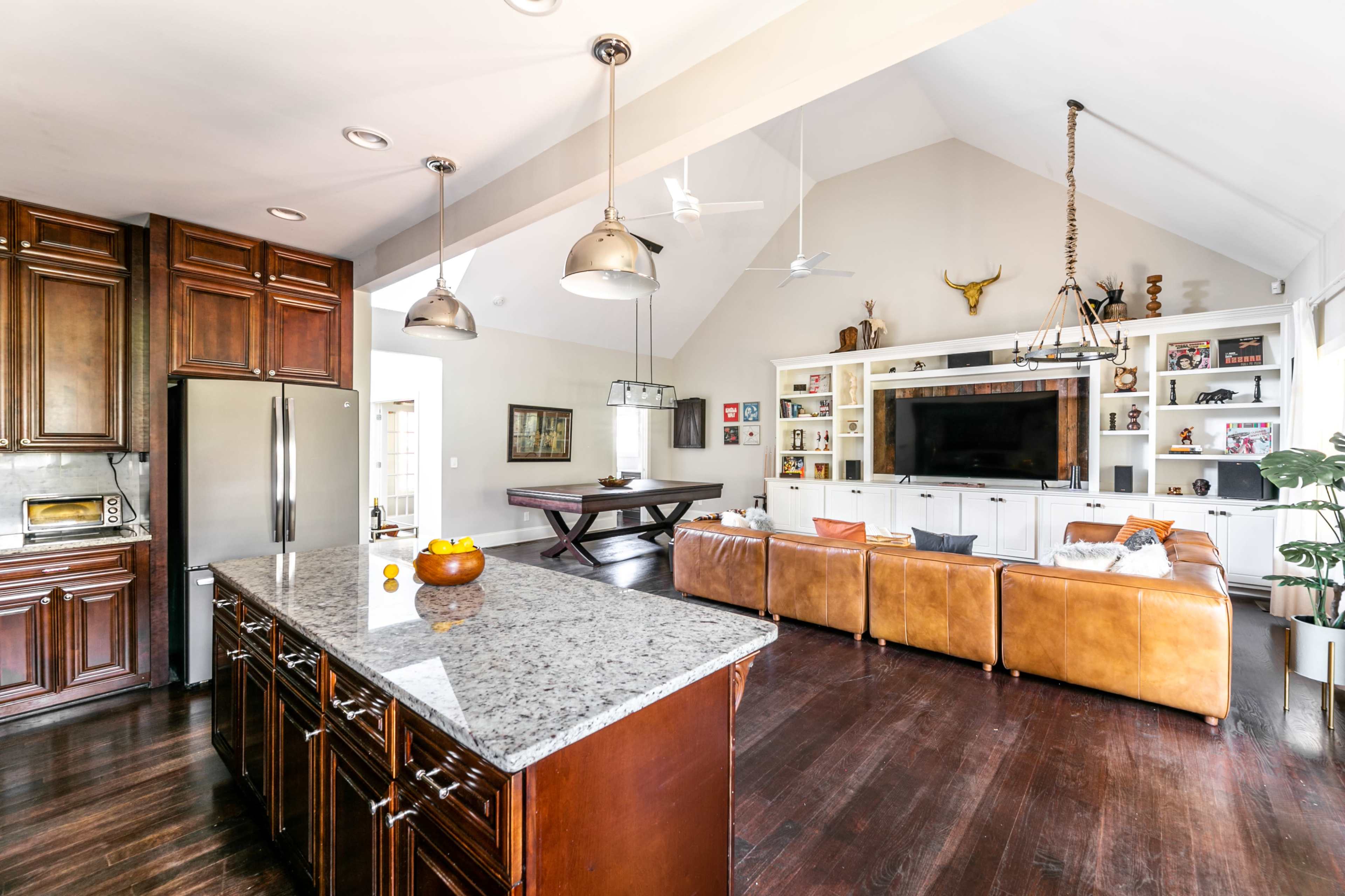 A spacious kitchen and living area features a granite island, wooden cabinetry, leather sofas, and a large entertainment unit.