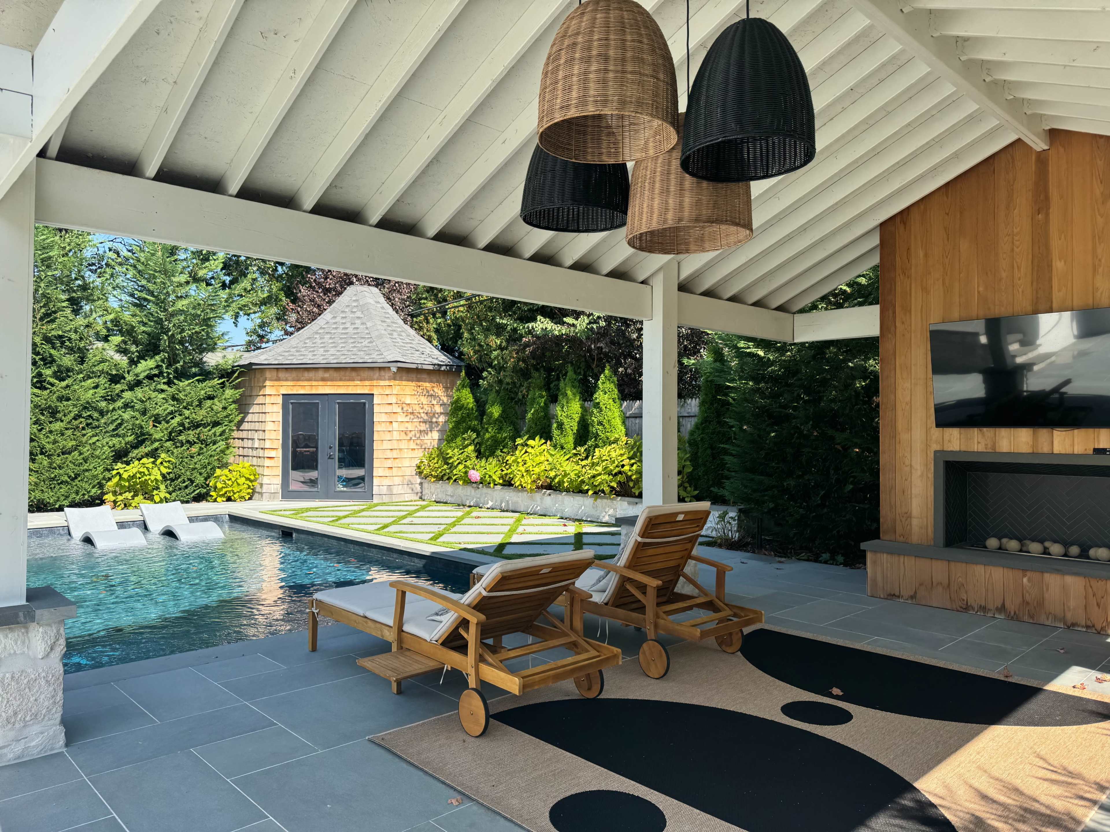The image shows a modern outdoor living space with two wooden lounge chairs, a swimming pool, and a small shed in a landscaped garden.