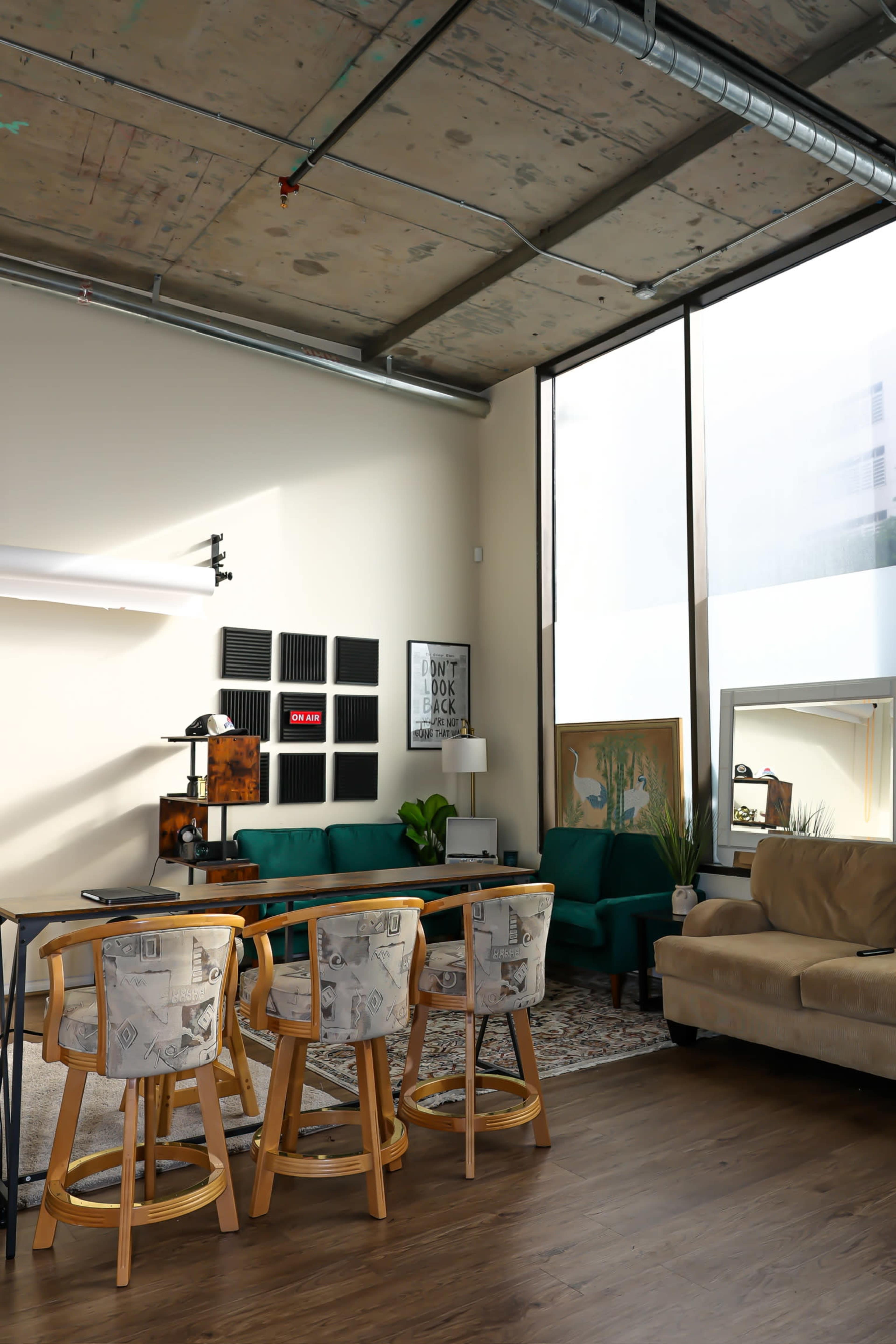 The image shows a modern living space with a large window, a green couch, a beige sofa, a dining table with four bar stools, and various decorative elements on the walls.