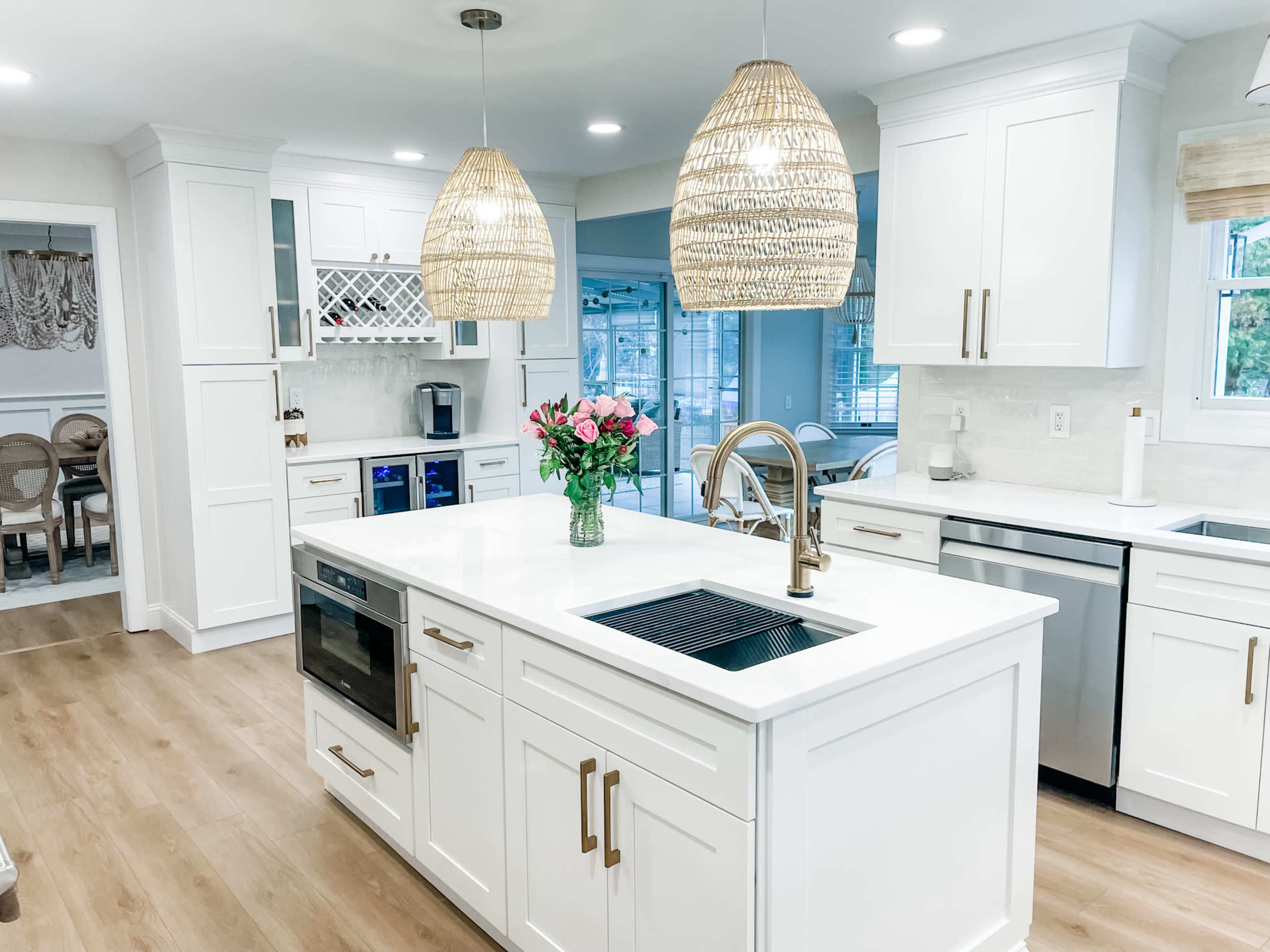 A modern kitchen features white cabinetry, an island with a sink, and pendant lighting above, with a vase of flowers on the countertop.