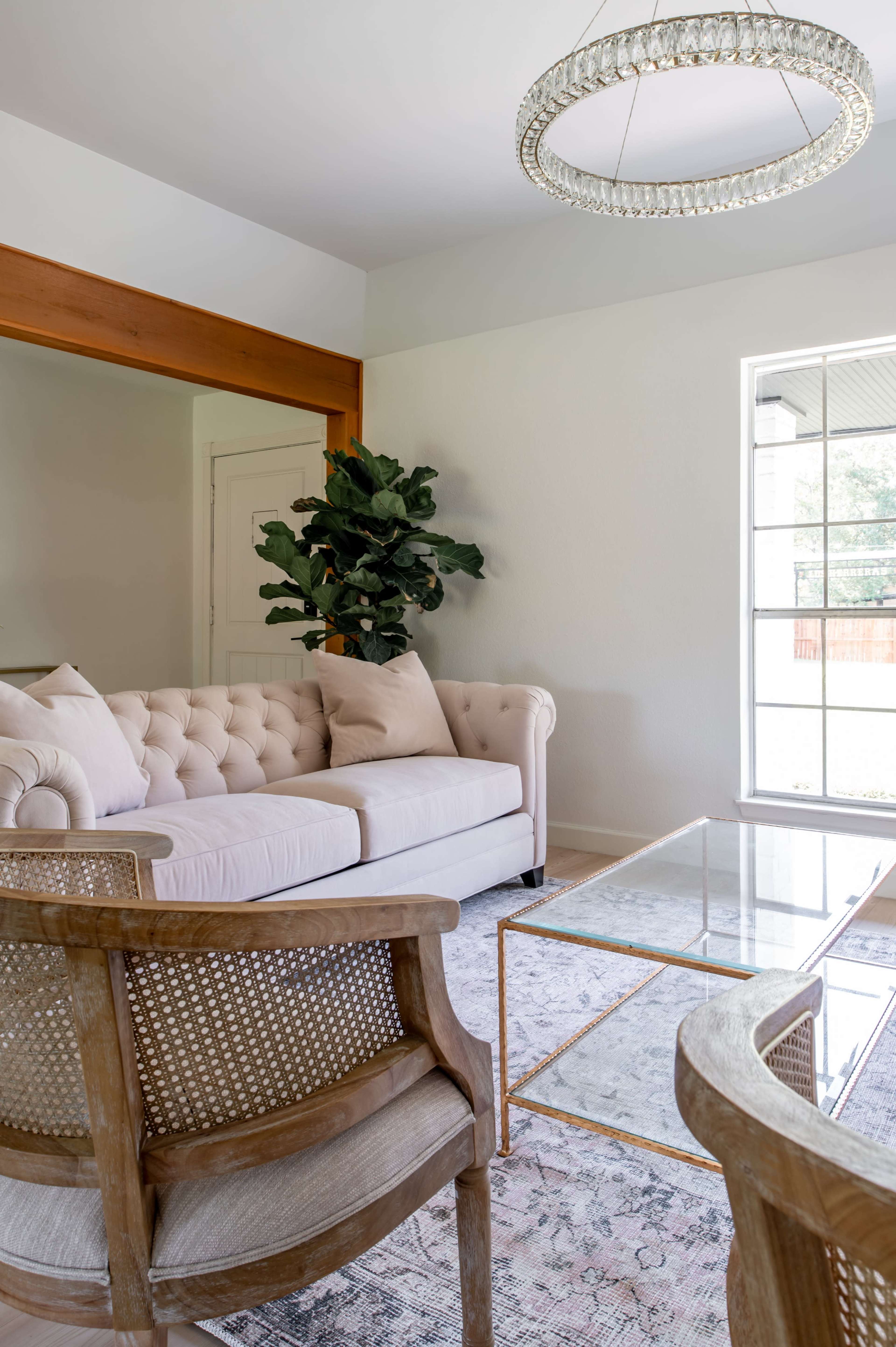 A light-colored tufted sofa and two wooden chairs are positioned around a glass coffee table in a well-lit living room with a potted plant and a large window.