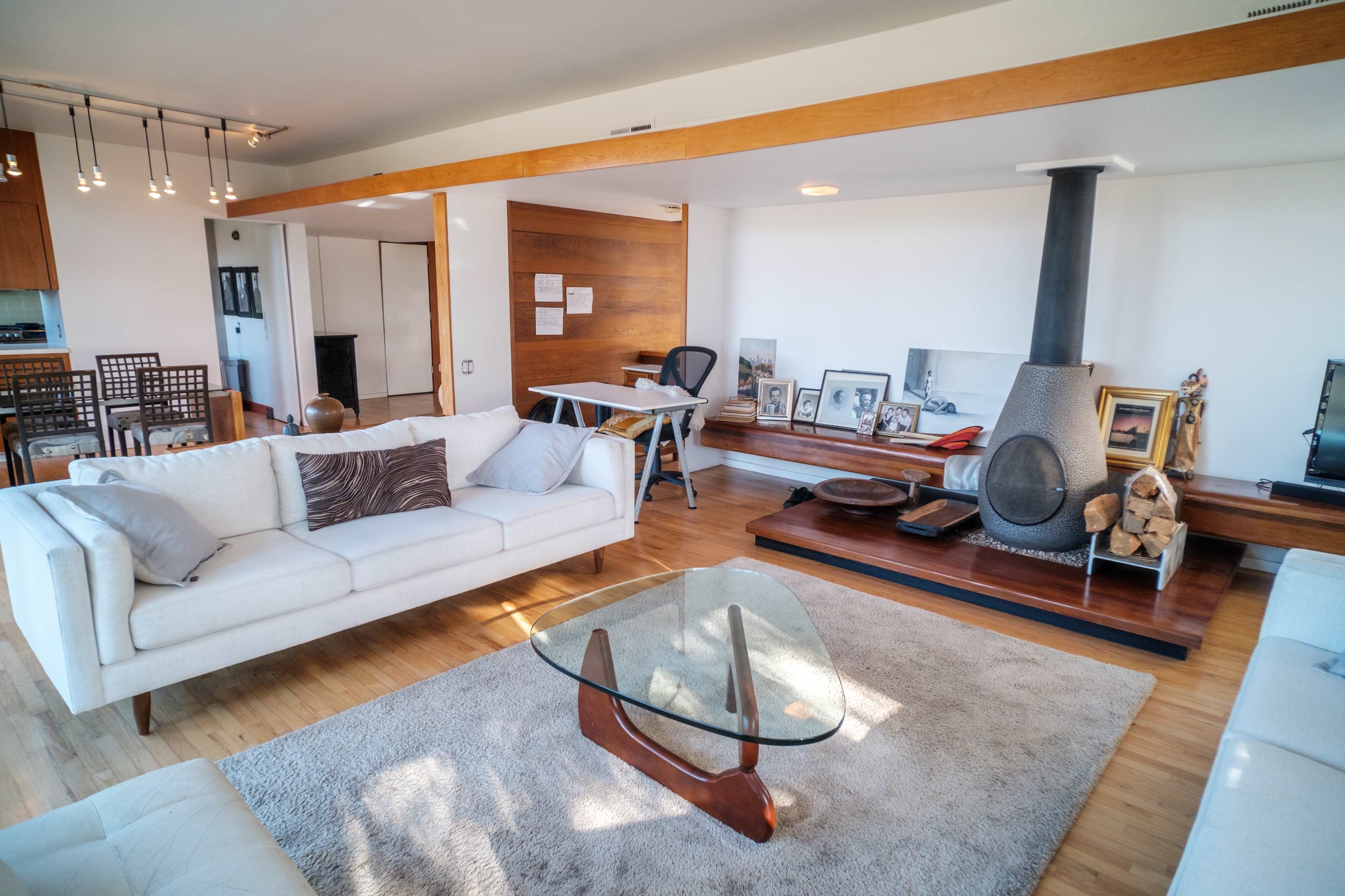 A modern living room with two white sofas facing a glass coffee table, a wood-burning stove, and a fireplace surrounded by wooden accents and various framed pictures on the walls.