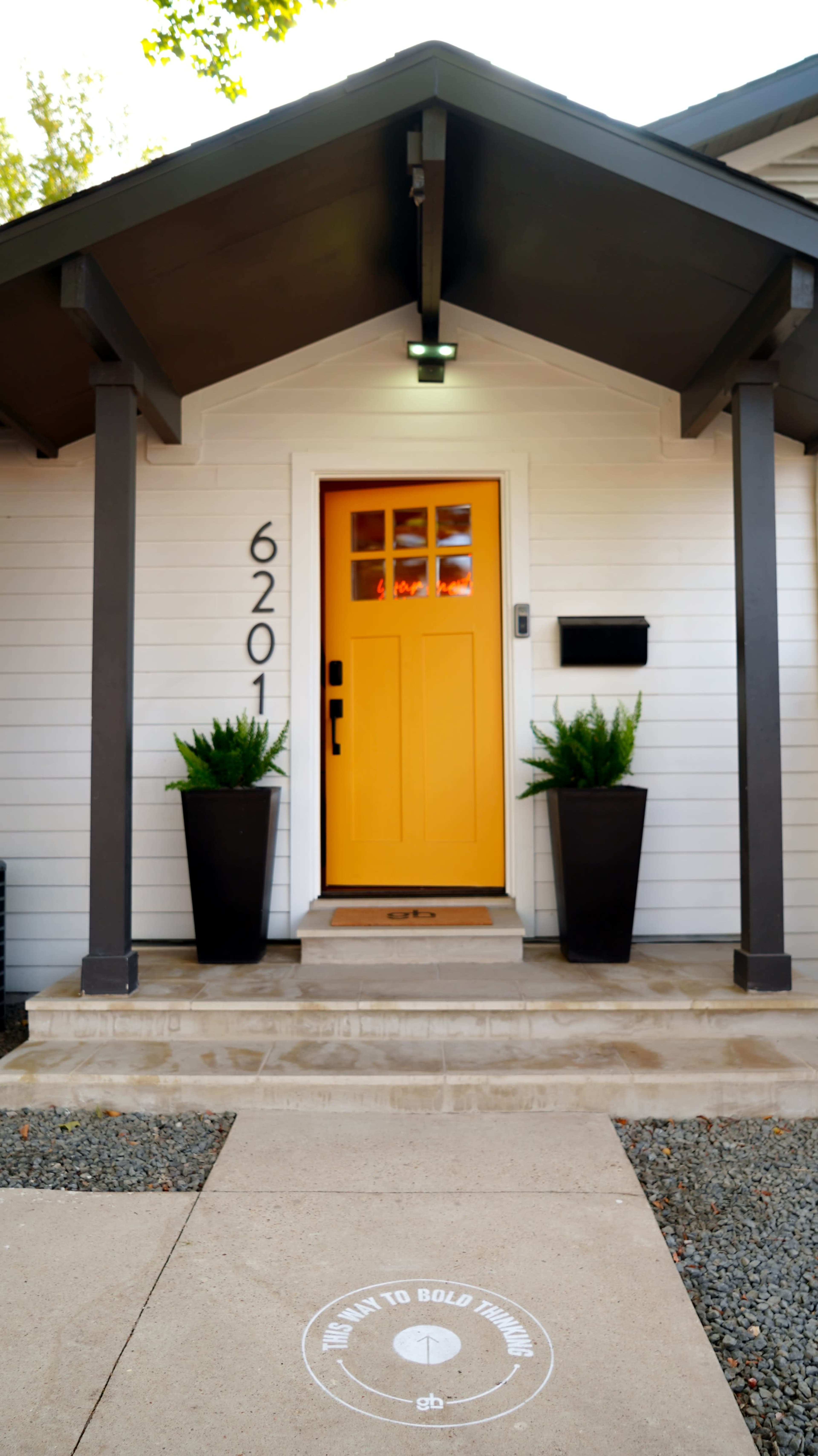 The image shows a front entrance of a house with a bright yellow door, flanked by two large planters with green plants, under a covered porch.