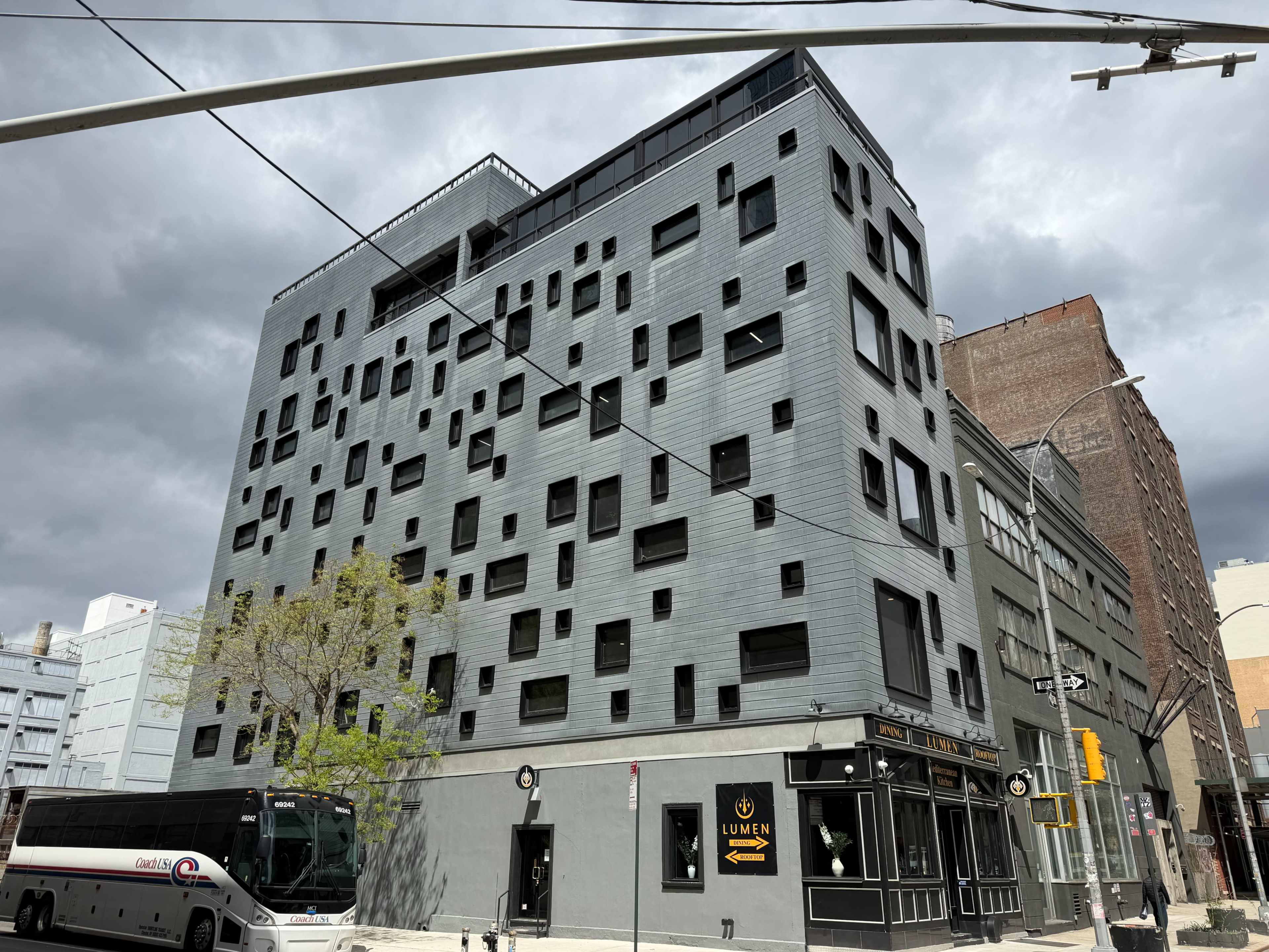A modern, gray apartment building featuring numerous square windows in varying sizes stands on a city street under an overcast sky.