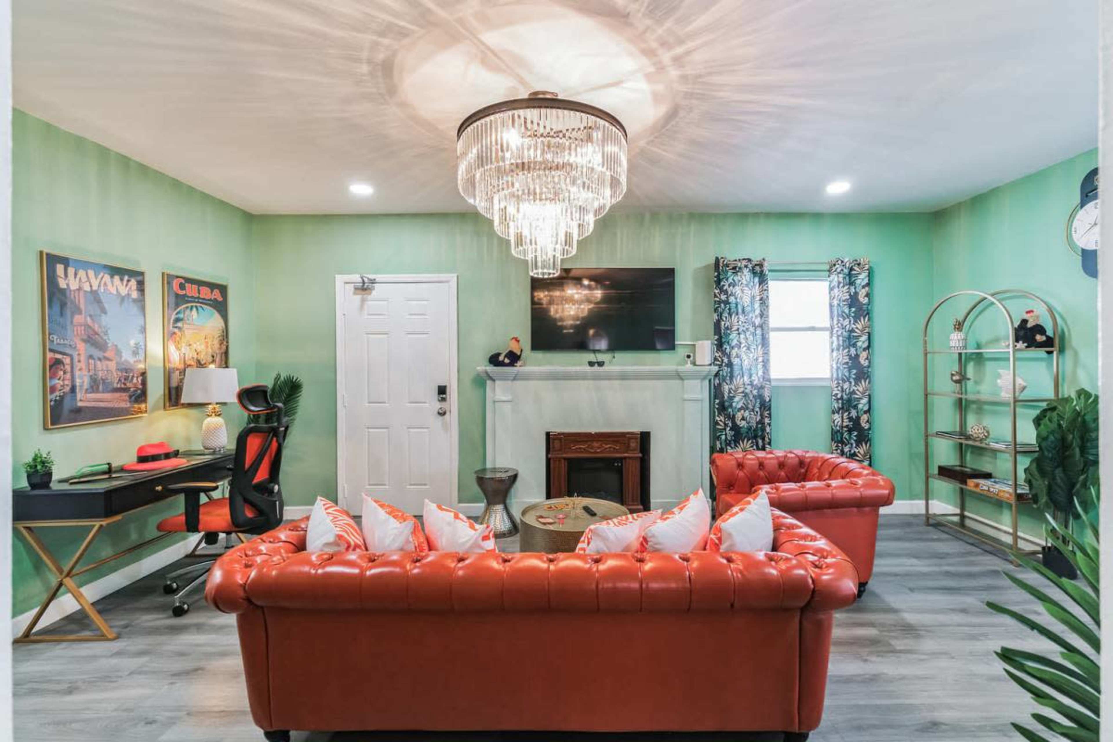 The image shows a brightly colored living room with a chandelier, a television above a fireplace, and a red leather sofa surrounded by decorative elements and artwork on the walls.