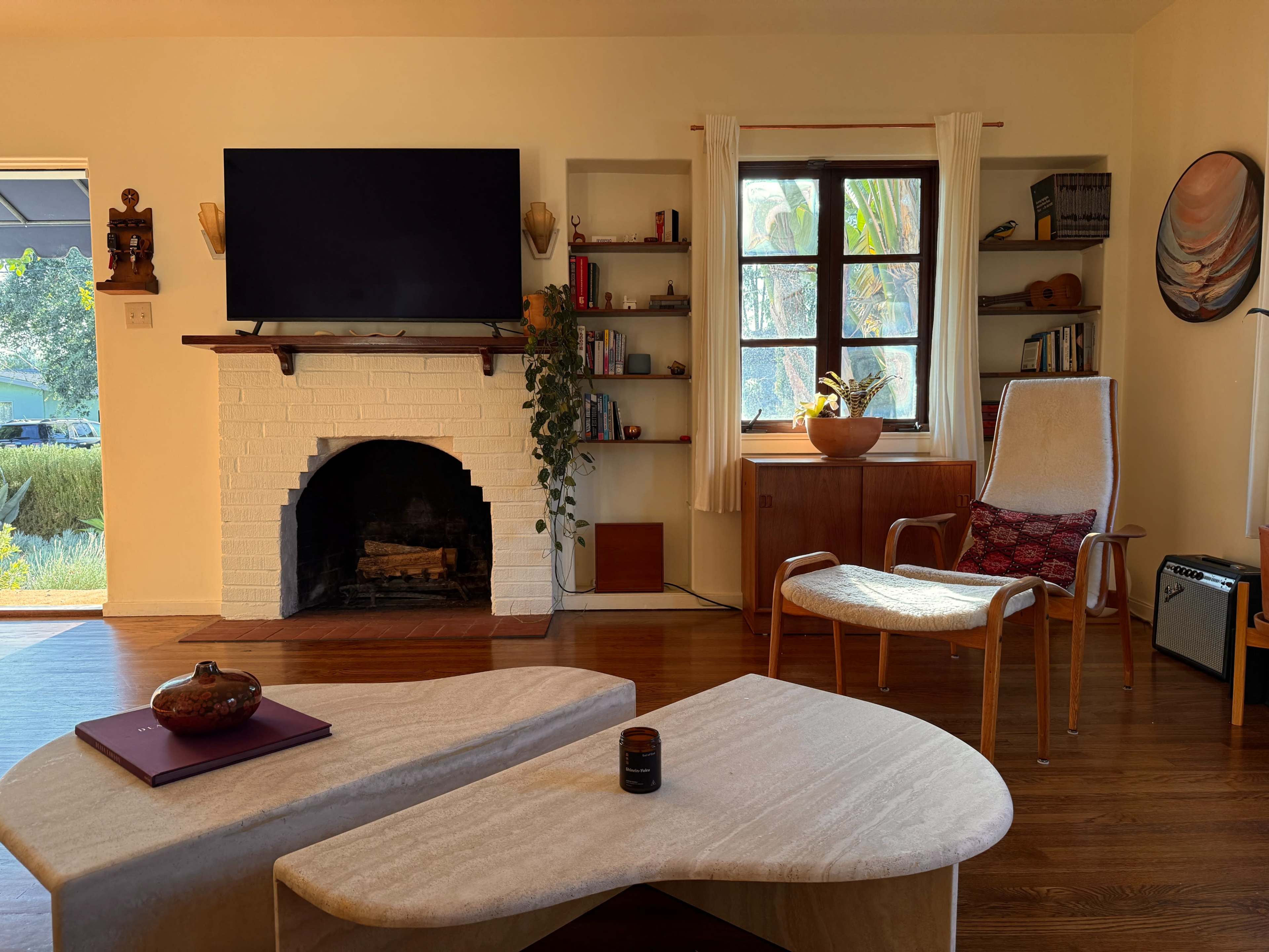 The image shows a modern living room featuring a white brick fireplace, a large TV mounted on the wall, a wooden coffee table, a chair with a soft cushion, and shelves with books and decor items.