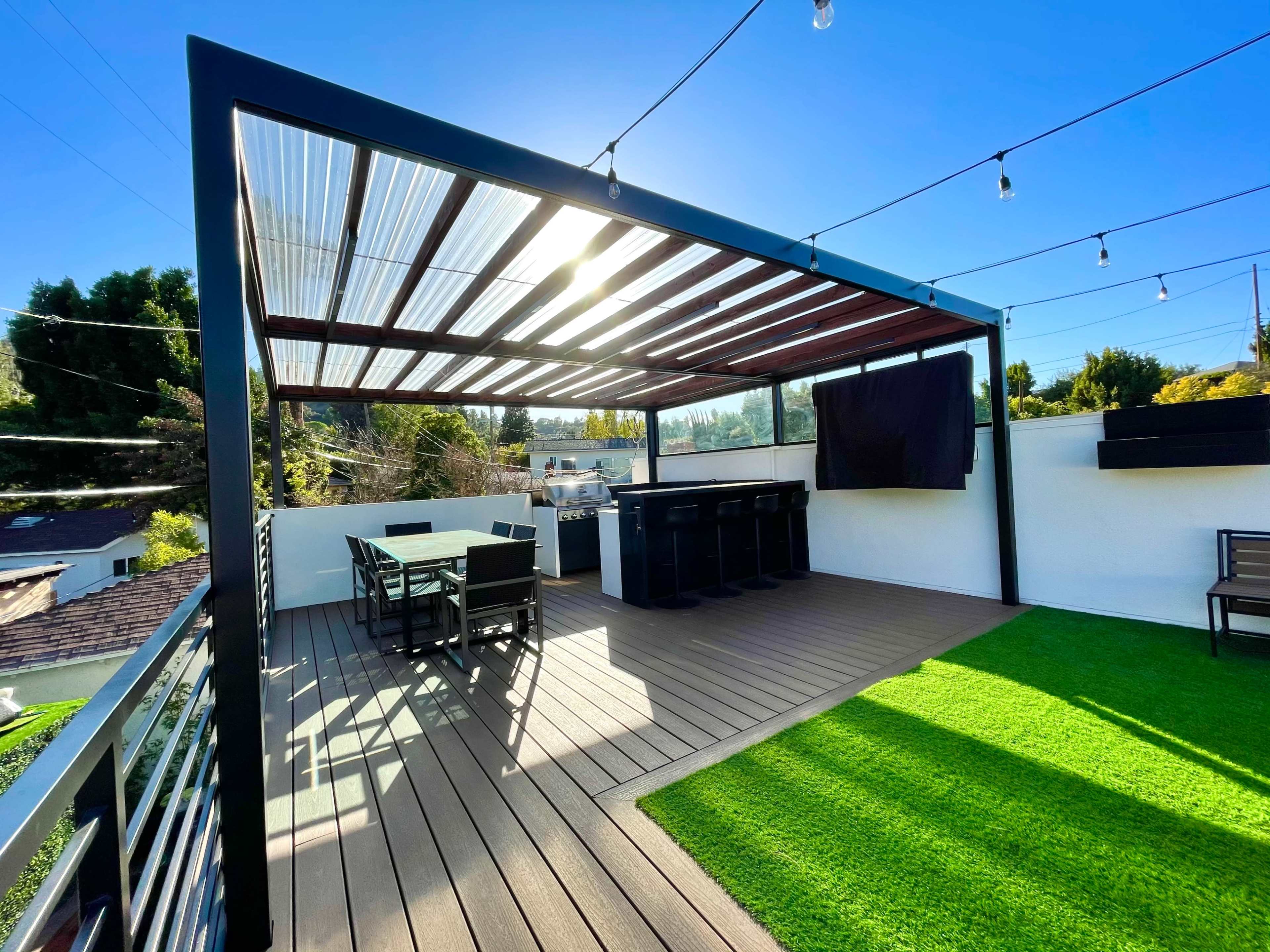 A modern outdoor patio features a wooden deck with a dining table and bar area, surrounded by greenery and string lights overhead.