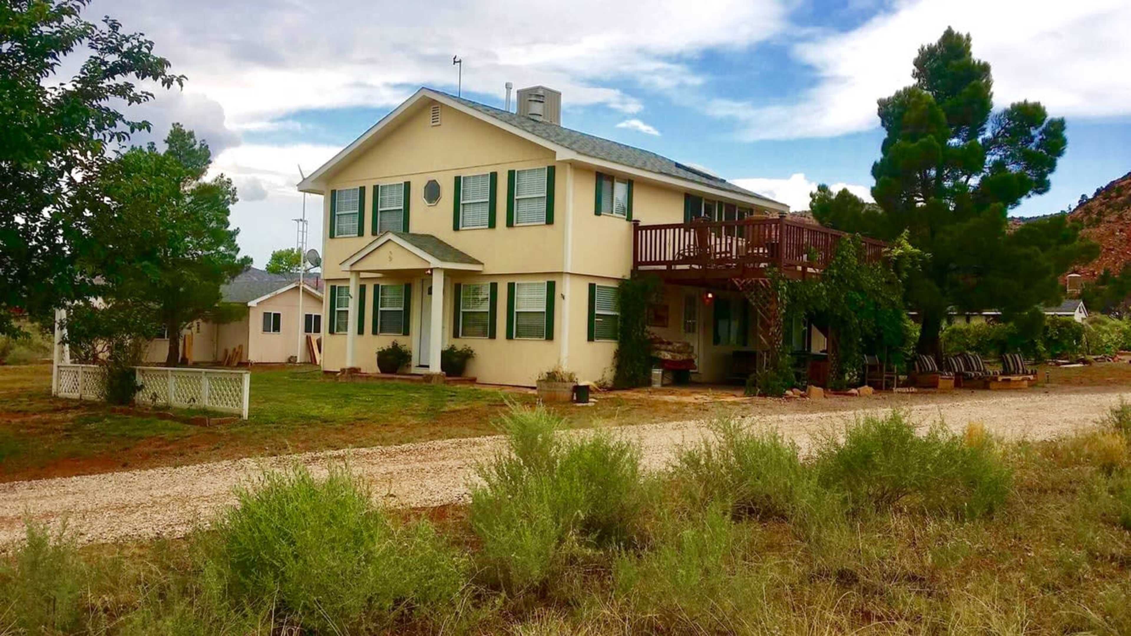 A two-story house with a balcony and surrounding greenery sits alongside a gravel driveway.