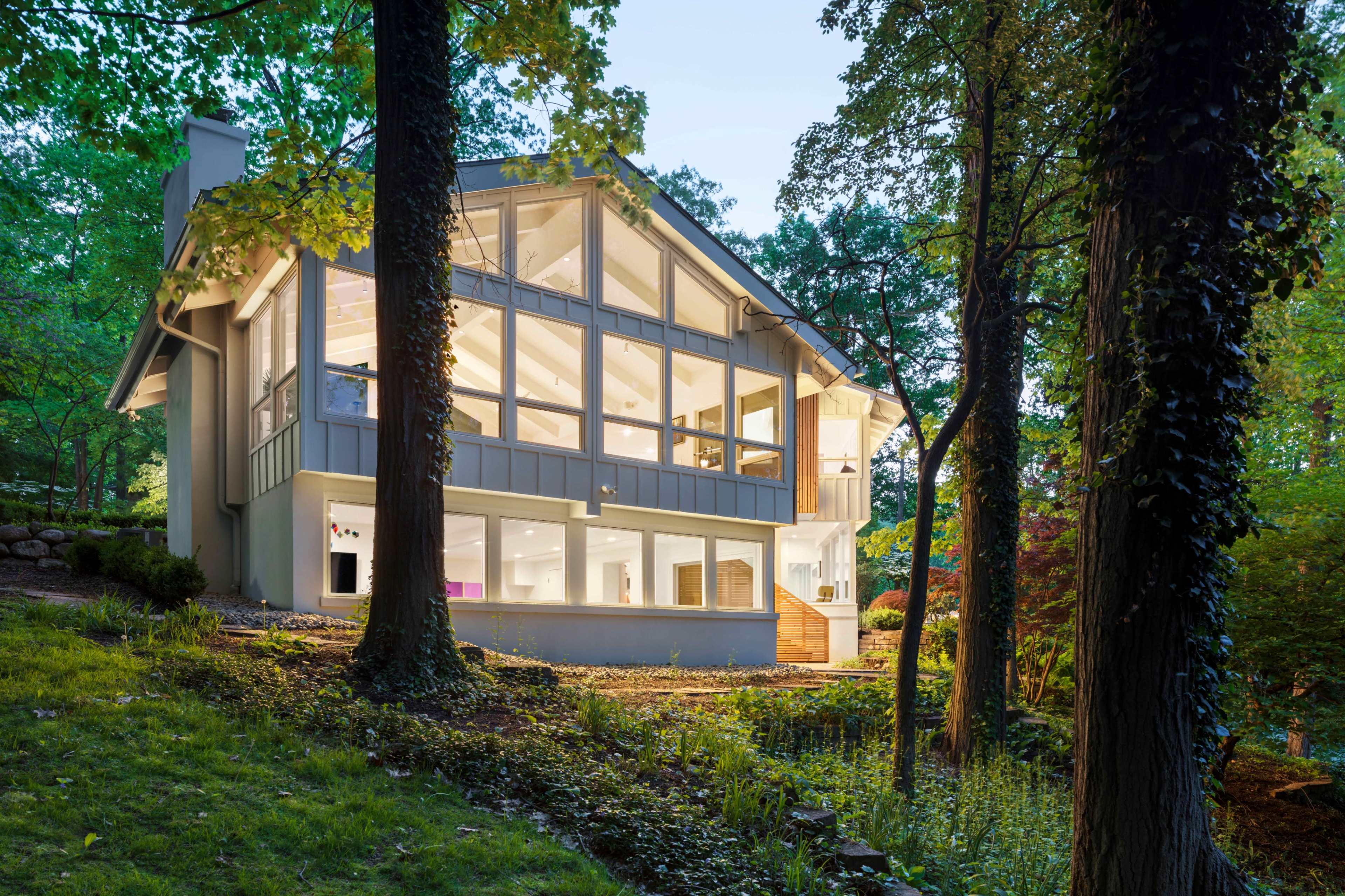 A modern house with large windows and a triangular roof is surrounded by trees in a wooded area during dusk.