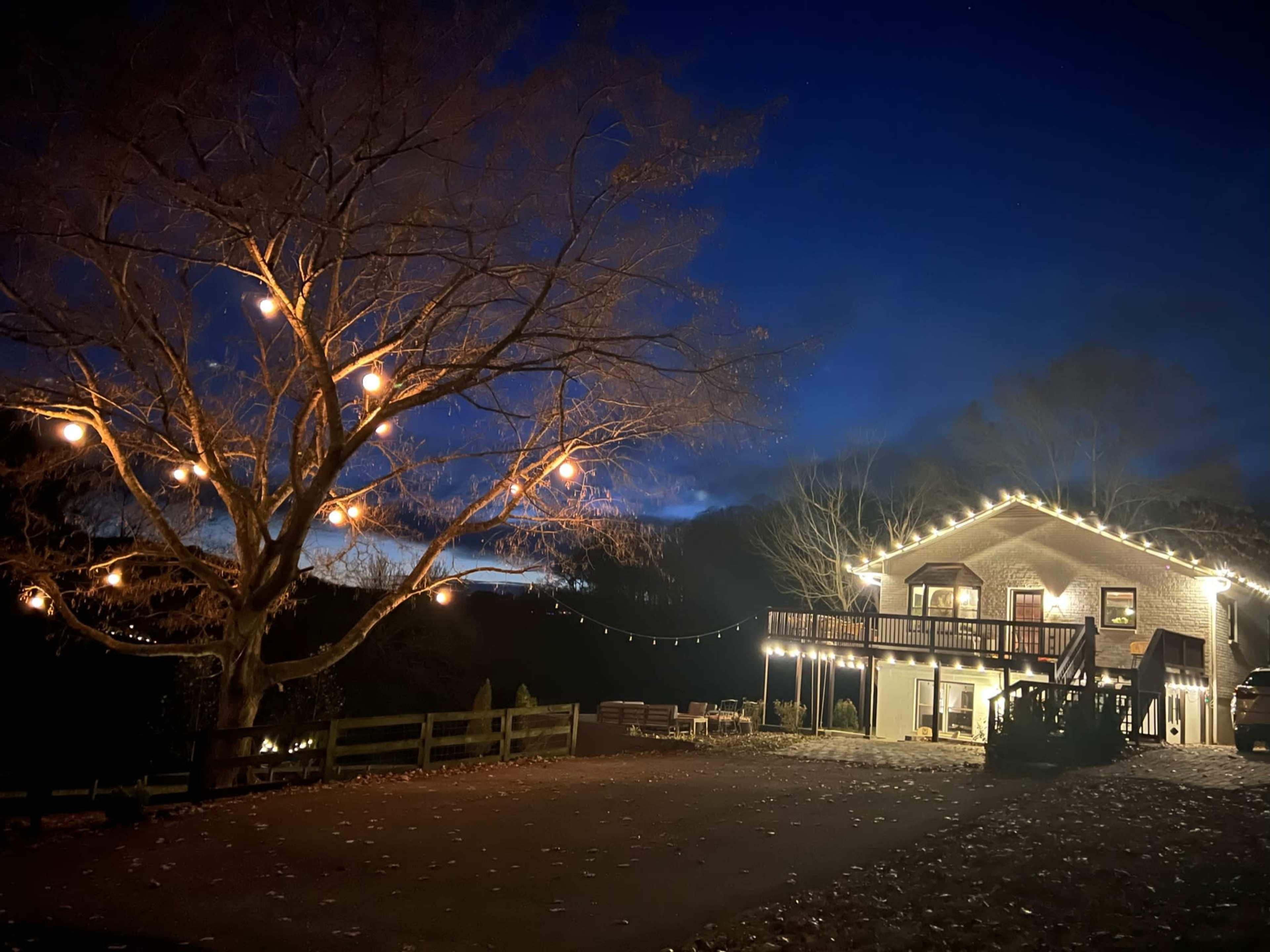 A decorated house with string lights stands beside a tree adorned with glowing orbs against a twilight sky.