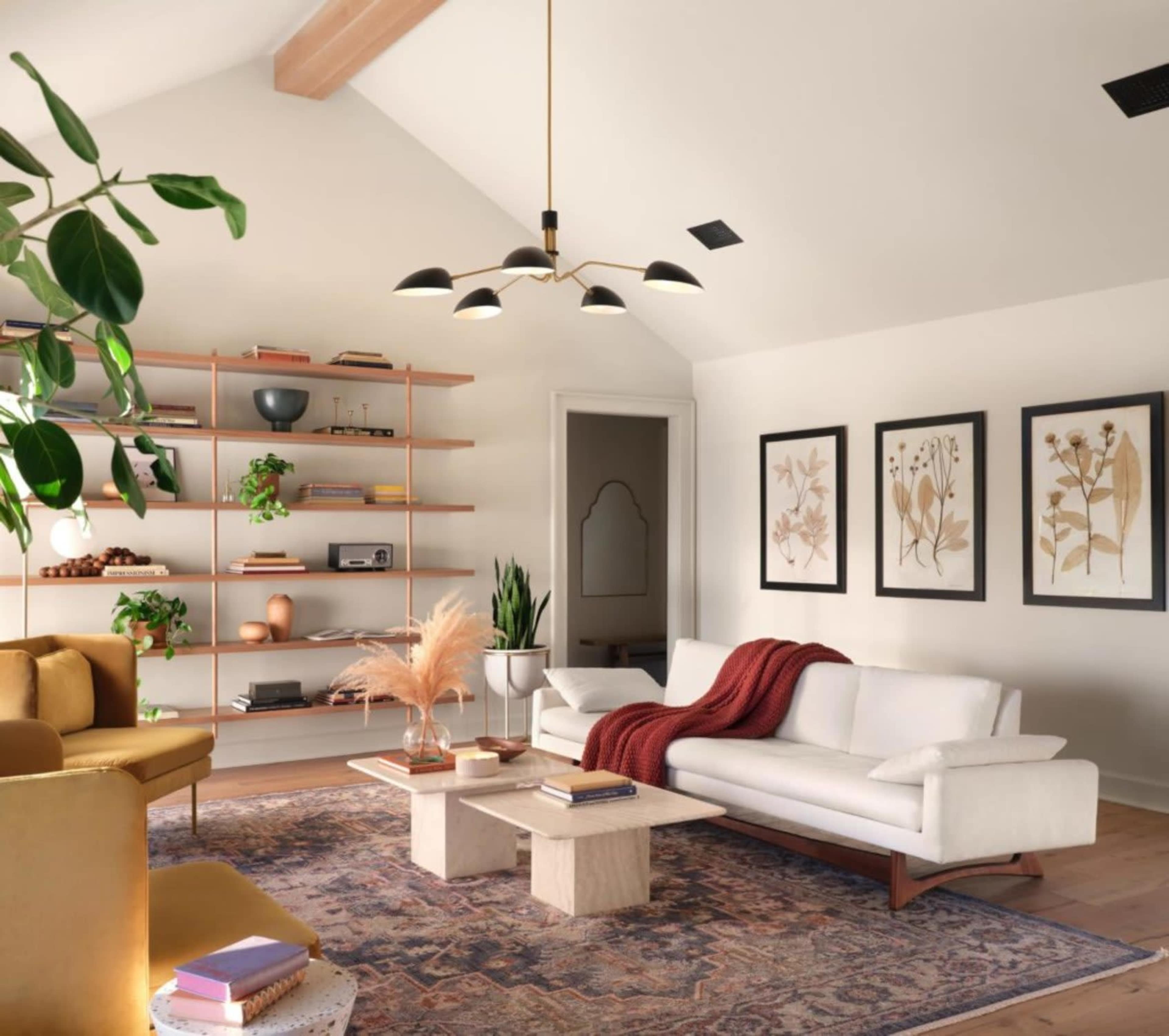 The room features a cozy living area with a white sofa, a mustard-colored armchair, a bookshelf filled with decor, and three framed botanical prints on the wall.