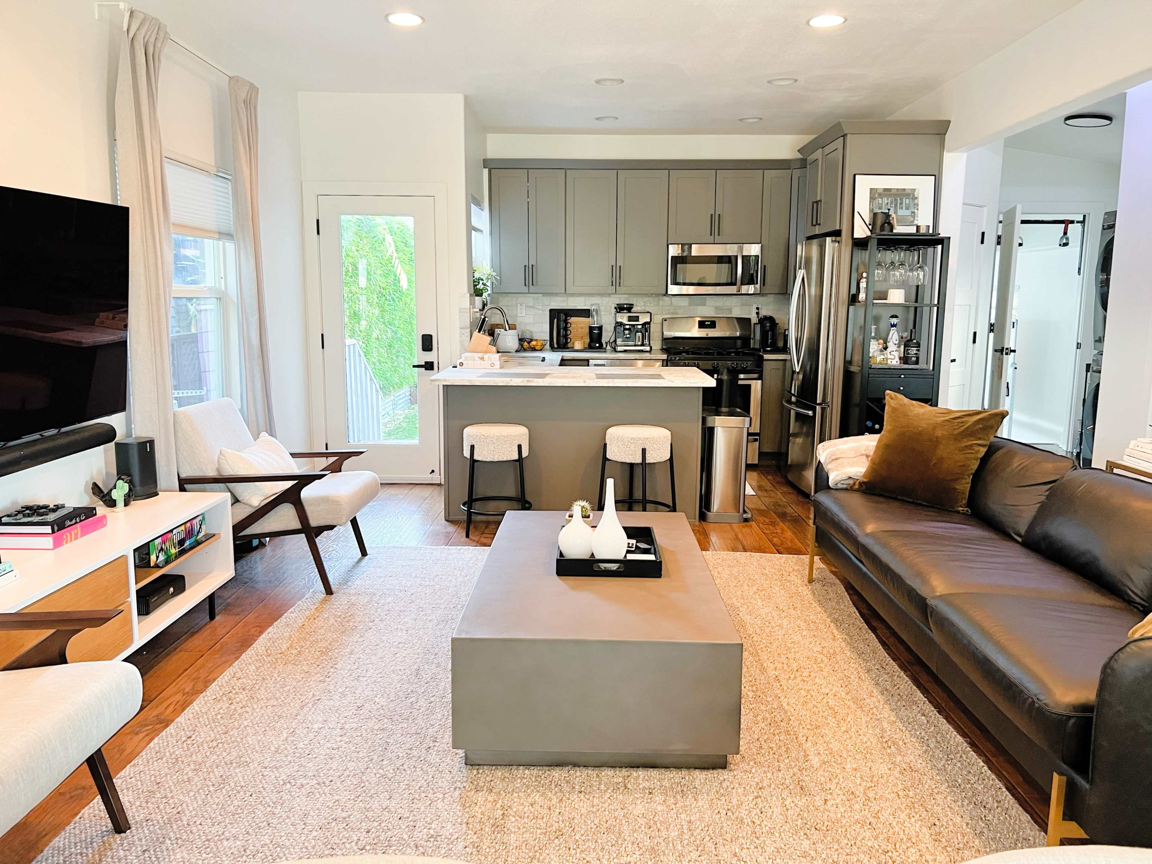 The image shows a modern living room and kitchen area featuring a leather sofa, a coffee table, and a kitchen with stainless steel appliances and grey cabinetry.