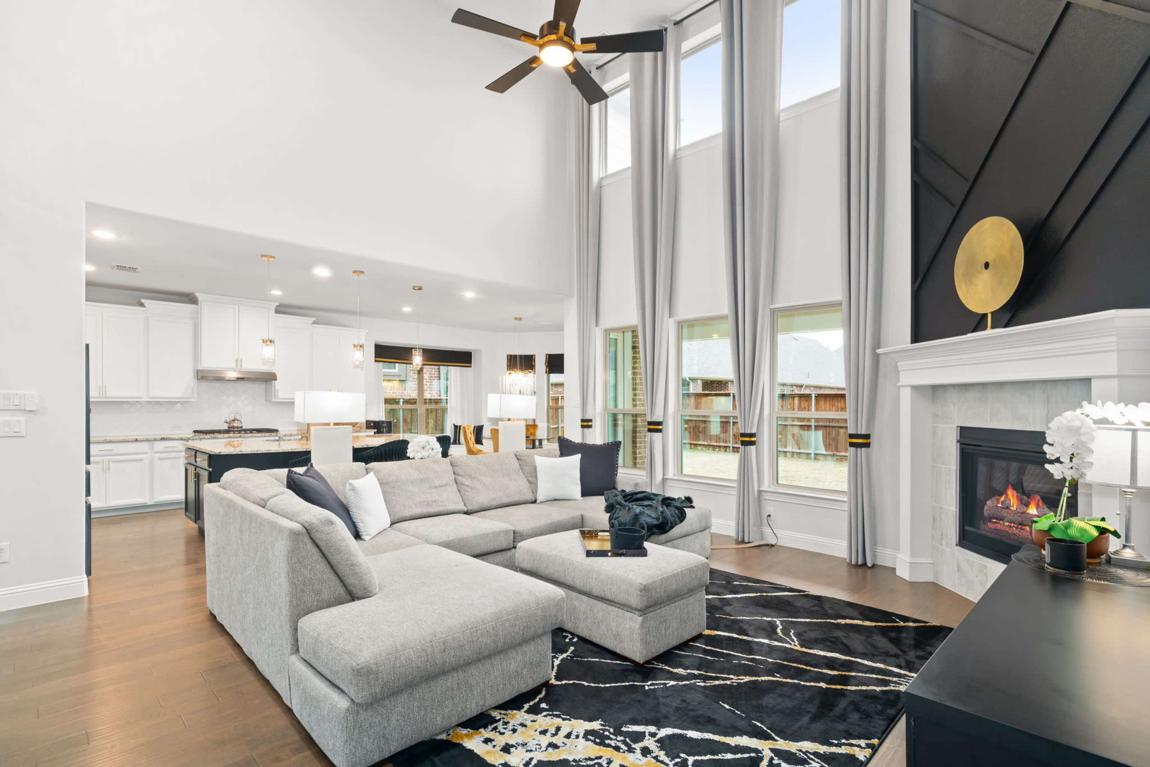 The image shows a modern living room featuring a large gray sectional sofa, a black and gold area rug, a fireplace, and tall windows allowing natural light to illuminate the space.