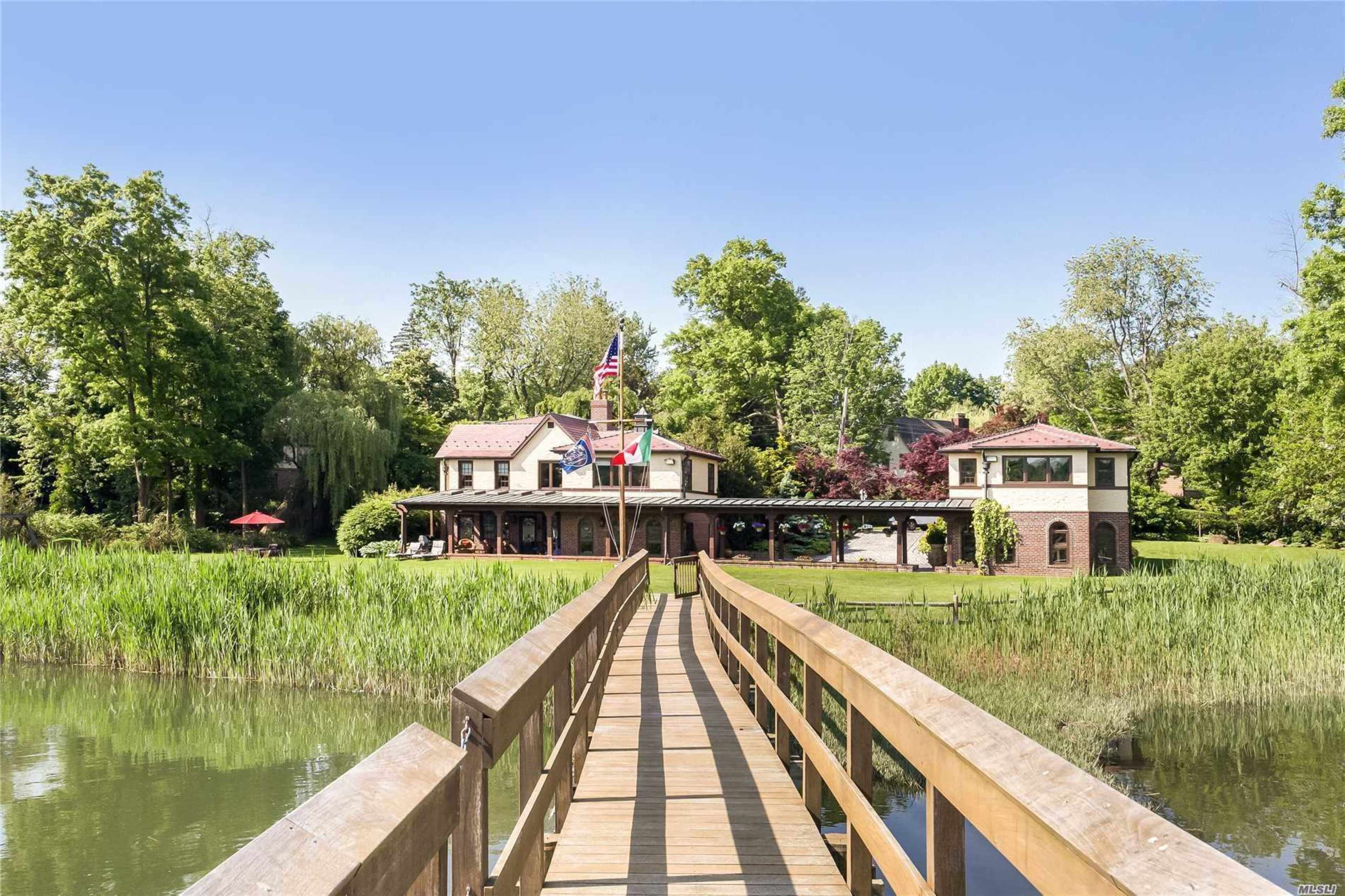 A wooden walkway leads to a large house surrounded by trees and greenery by a calm body of water.