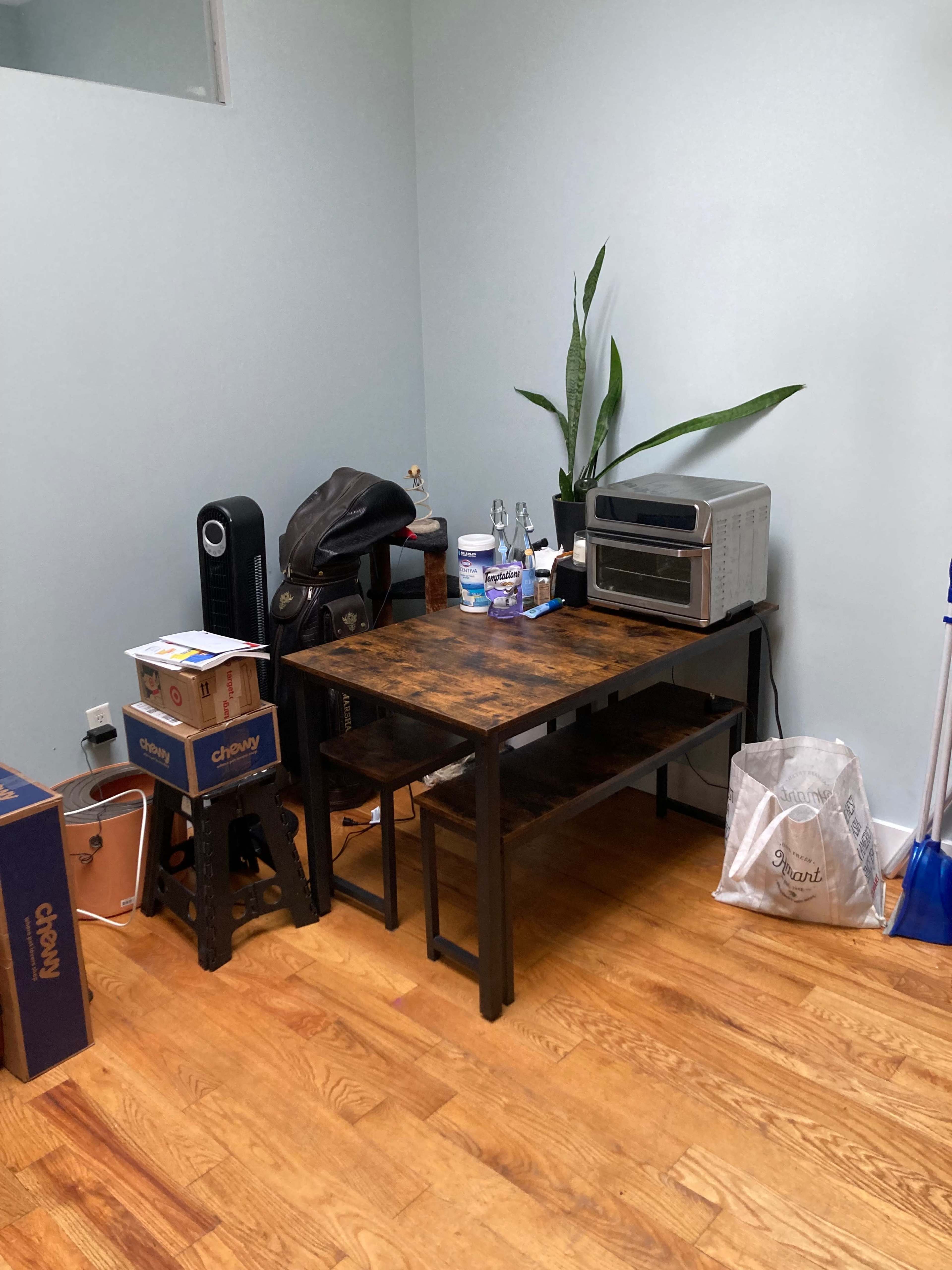 A wooden table with two benches is positioned in a corner of a room, accompanied by a small indoor plant, kitchen appliances, and various items on the floor.