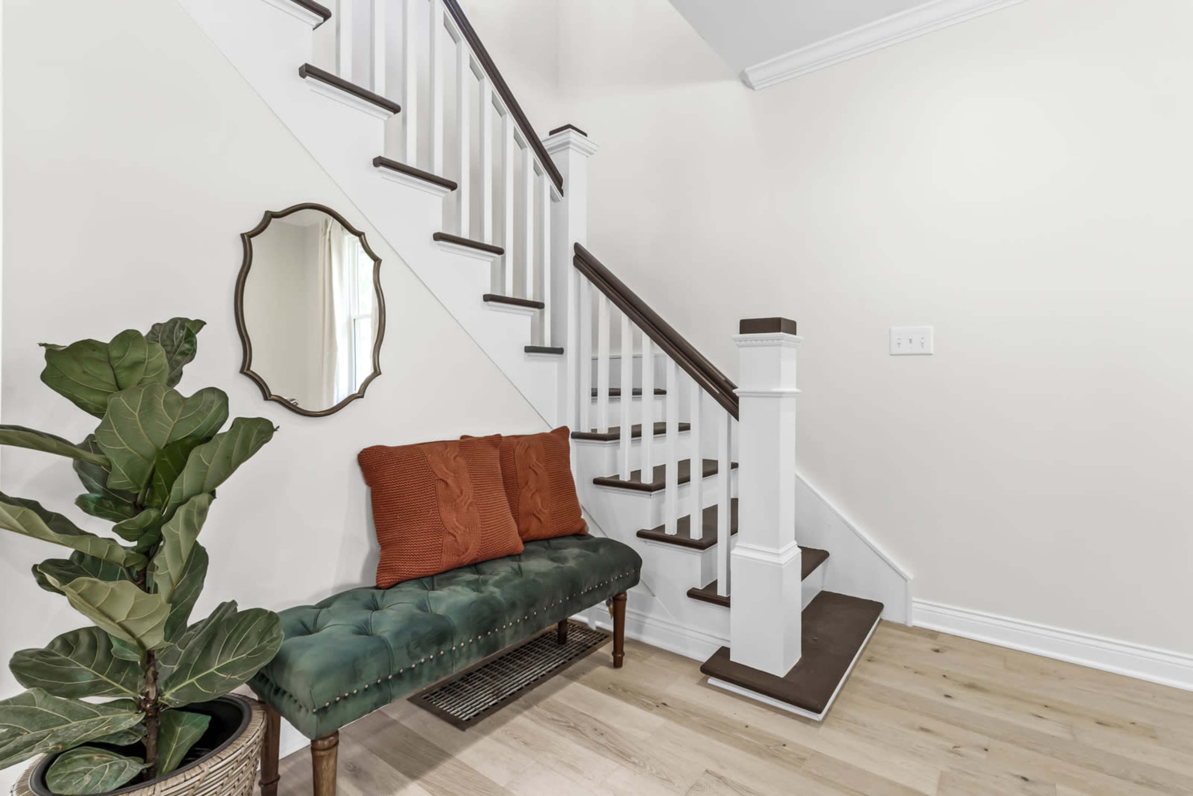 A staircase in a hallway with a green upholstered bench, a round mirror, and a potted plant against a light-colored wall.
