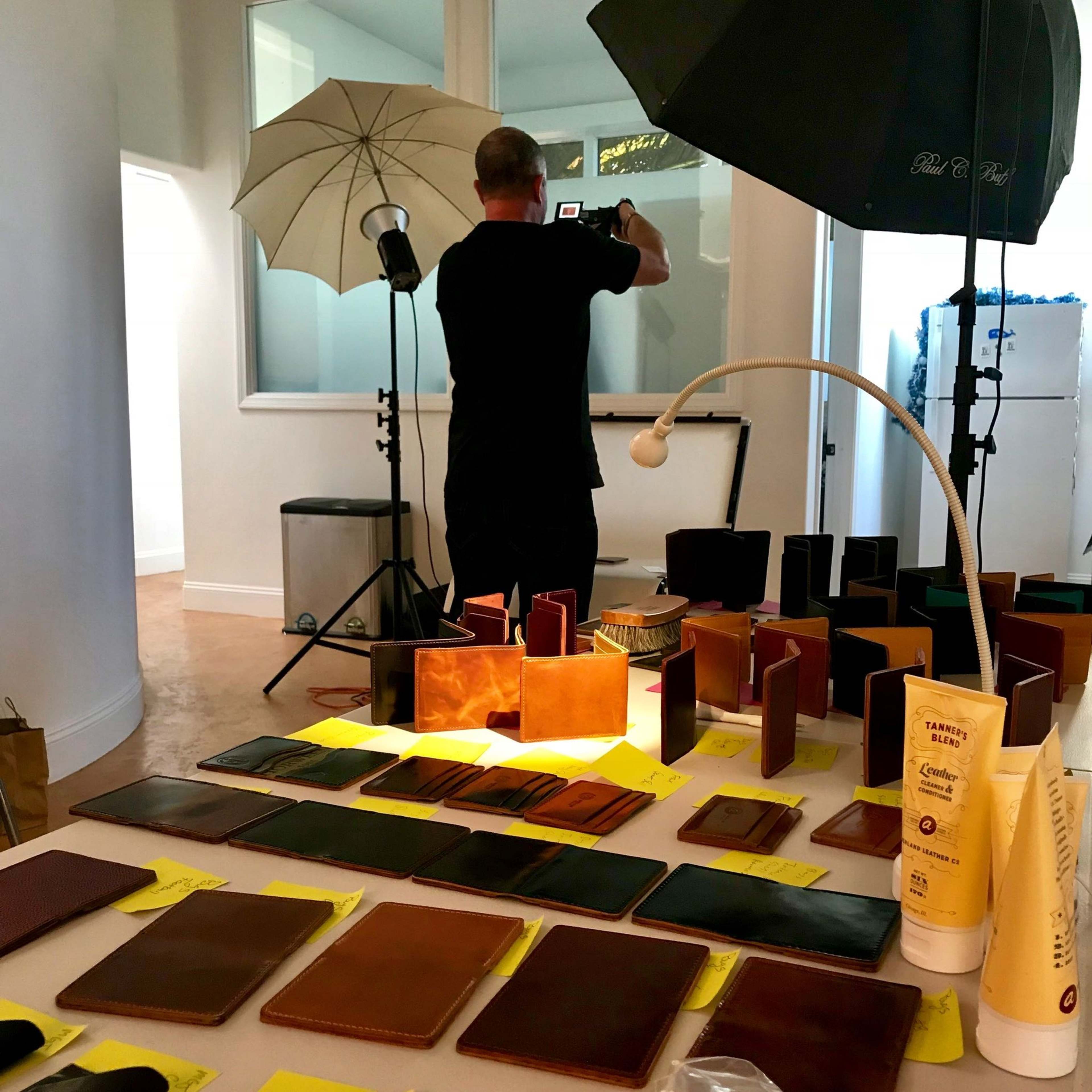 A person is photographing a display of leather goods arranged on a table, with studio lighting in the background.