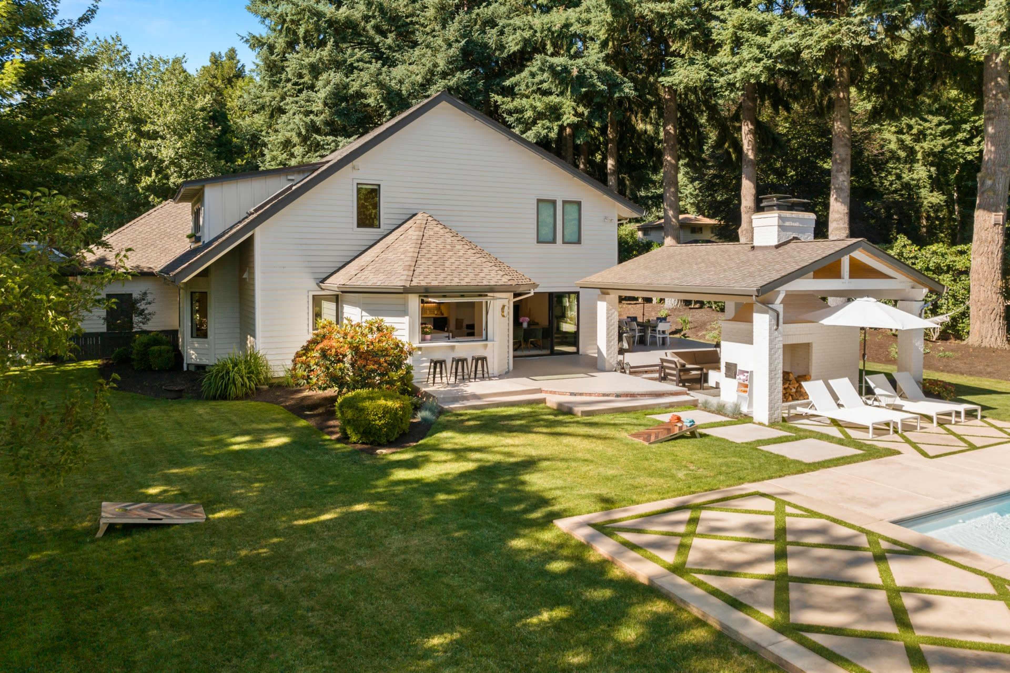 A two-story white house with a patio and outdoor seating area, surrounded by lush greenery and a swimming pool.