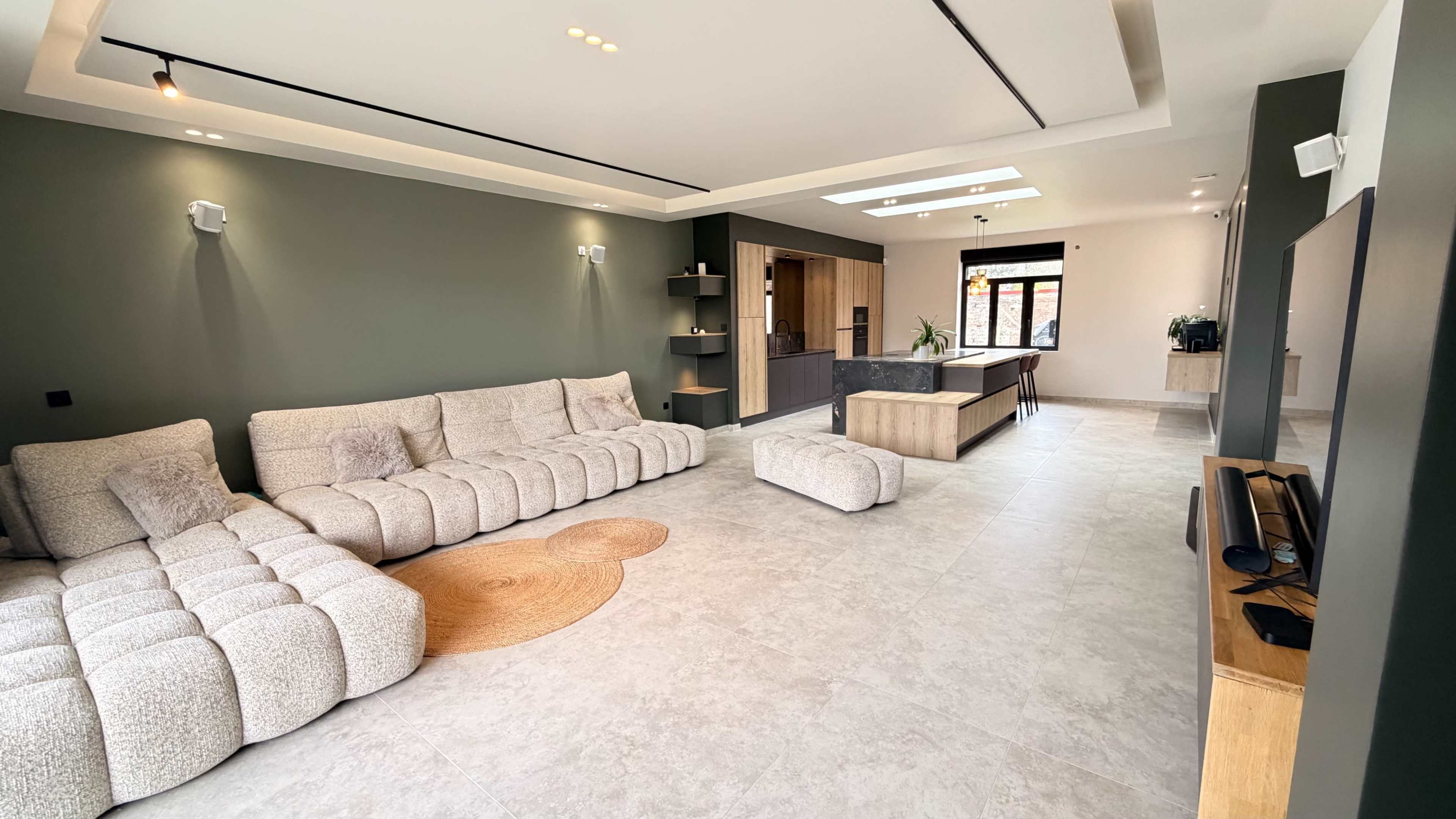 The image shows a modern living room featuring a large, plush sectional sofa, a circular area rug, and an open kitchen with a minimalist design.