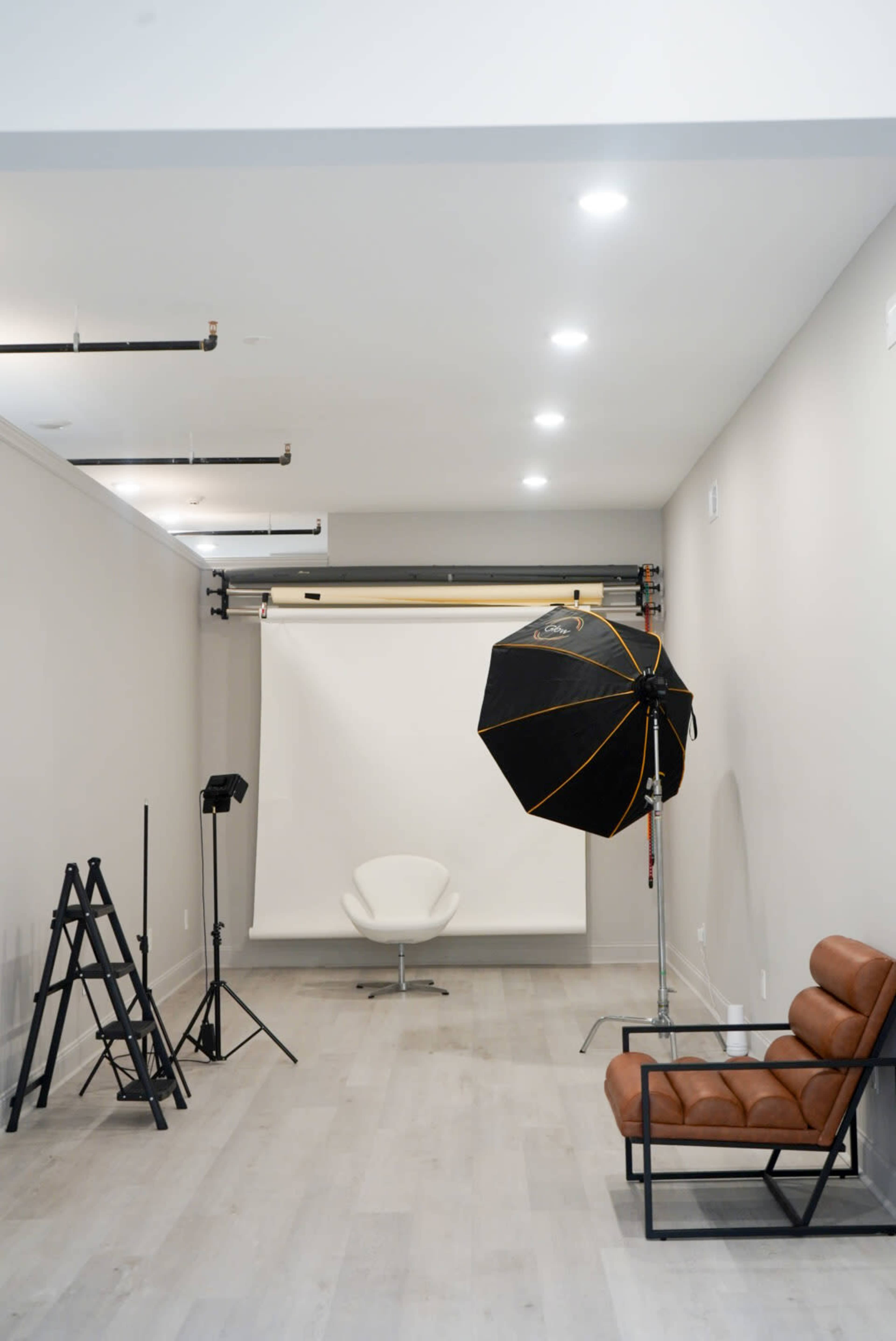The image shows a minimalist studio setup with a lighting umbrella, camera tripod, and a white backdrop with a chair in front, along with a brown leather sofa and a ladder against the wall.