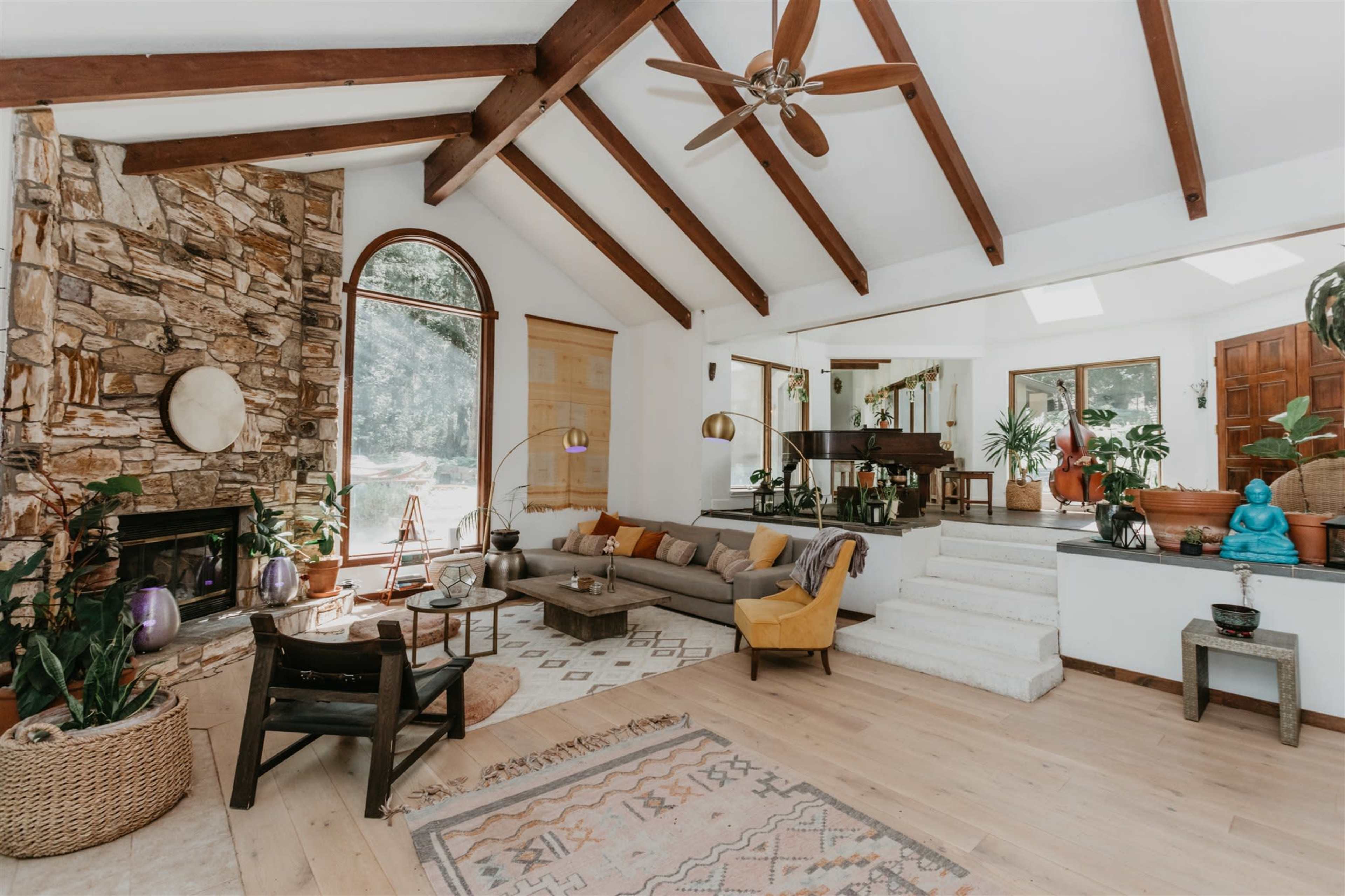 A spacious living room with a high ceiling, wooden beams, a stone fireplace, and various plants scattered throughout the area.