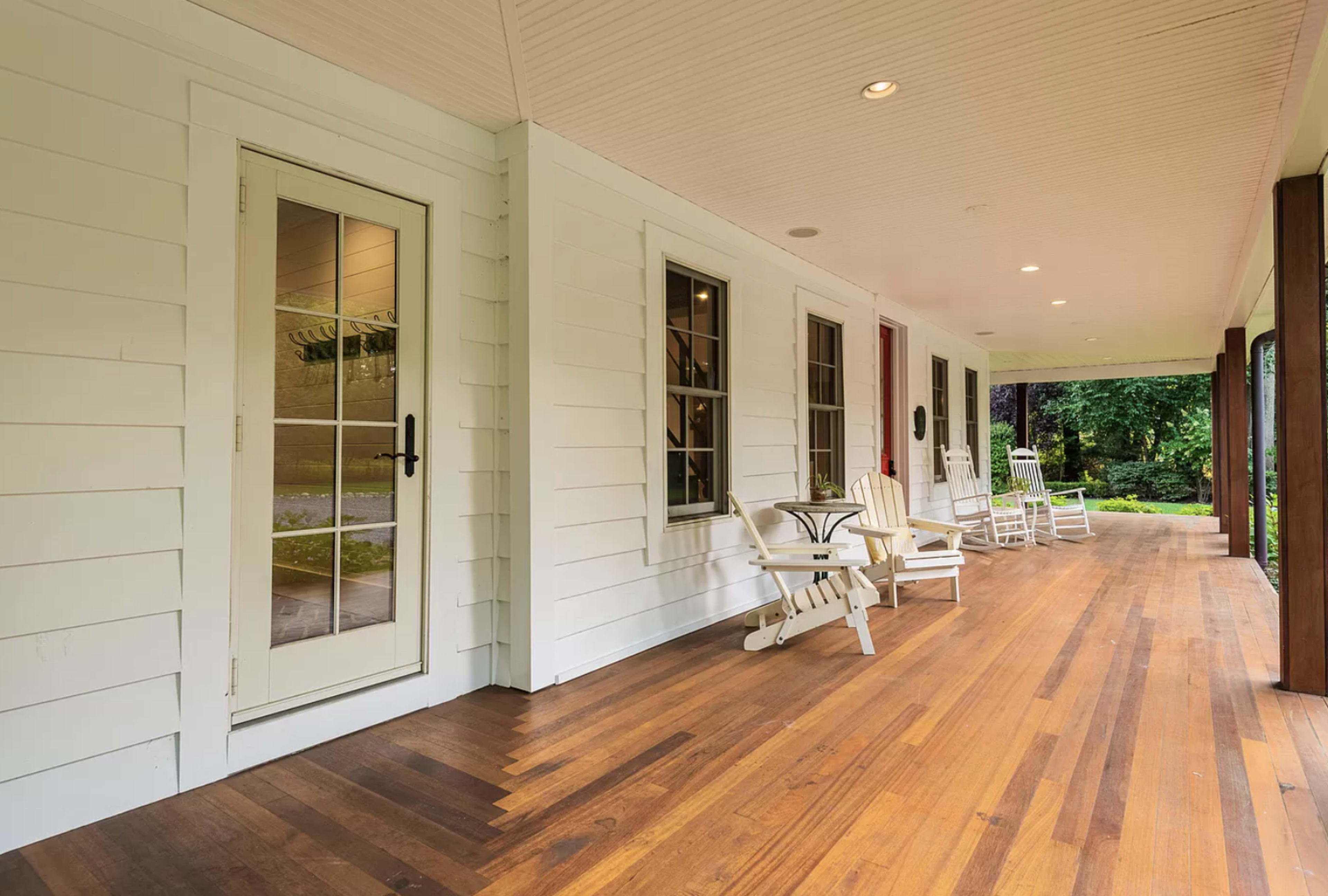 The image shows a porch with wooden flooring, featuring a white door, several rocking chairs, and a serene outdoor view.