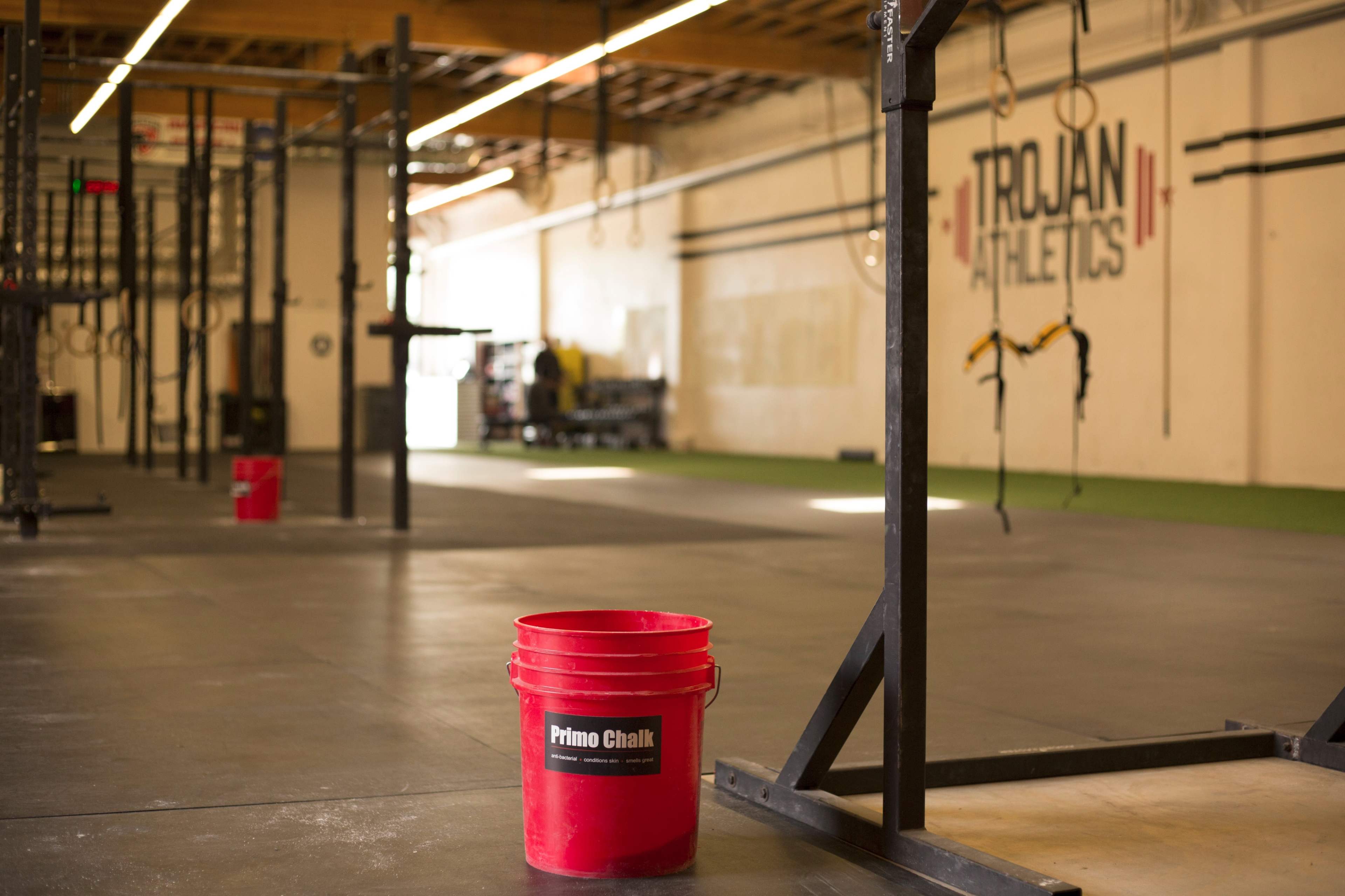 The image shows an empty gym space with workout equipment in the background and a red bucket labeled "Primo Chalk" in the foreground.