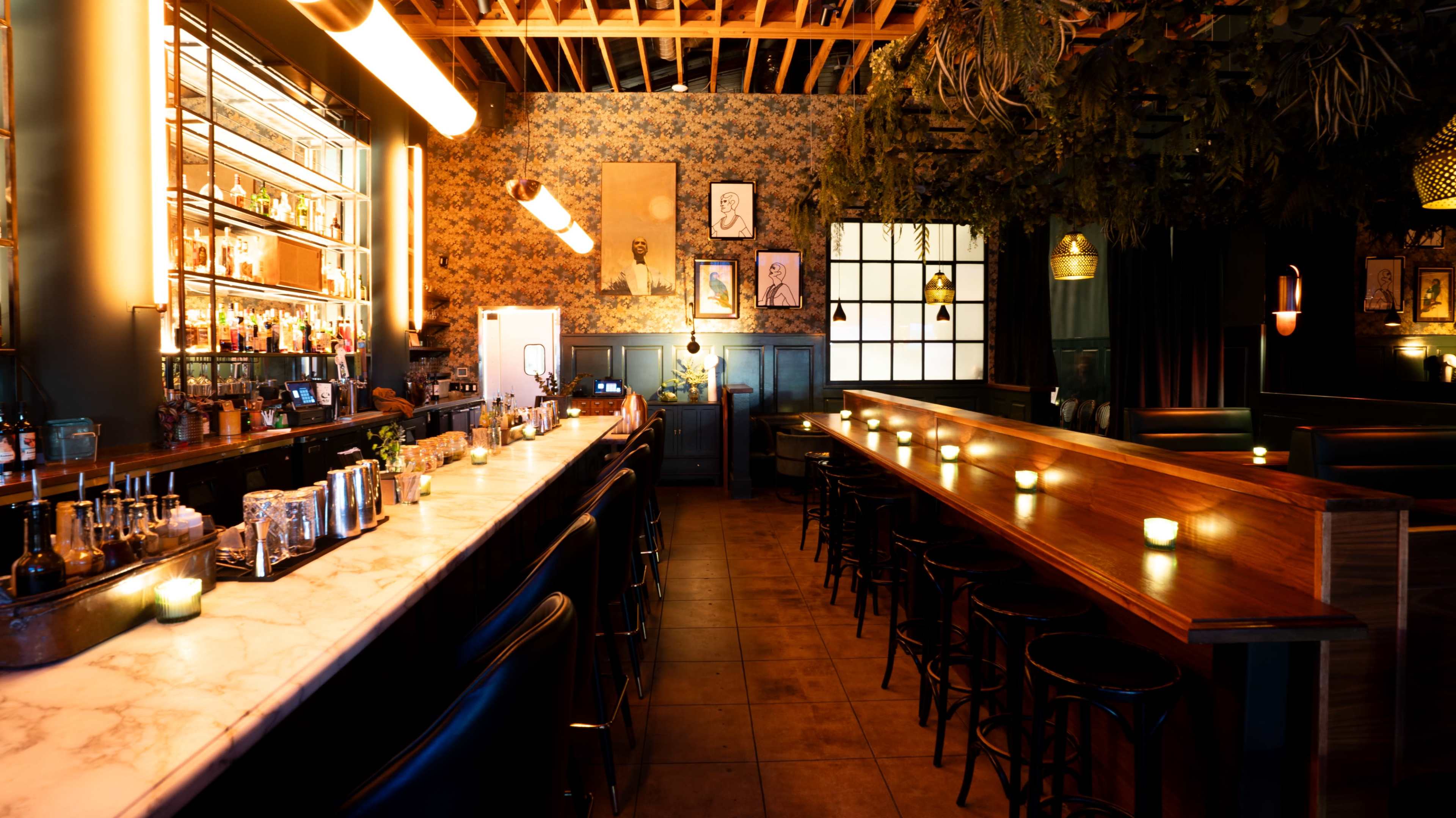 The image shows an elegantly designed bar interior with a long marble countertop, high stools, and ambient lighting, complemented by floral wallpaper and decorative elements on the walls.