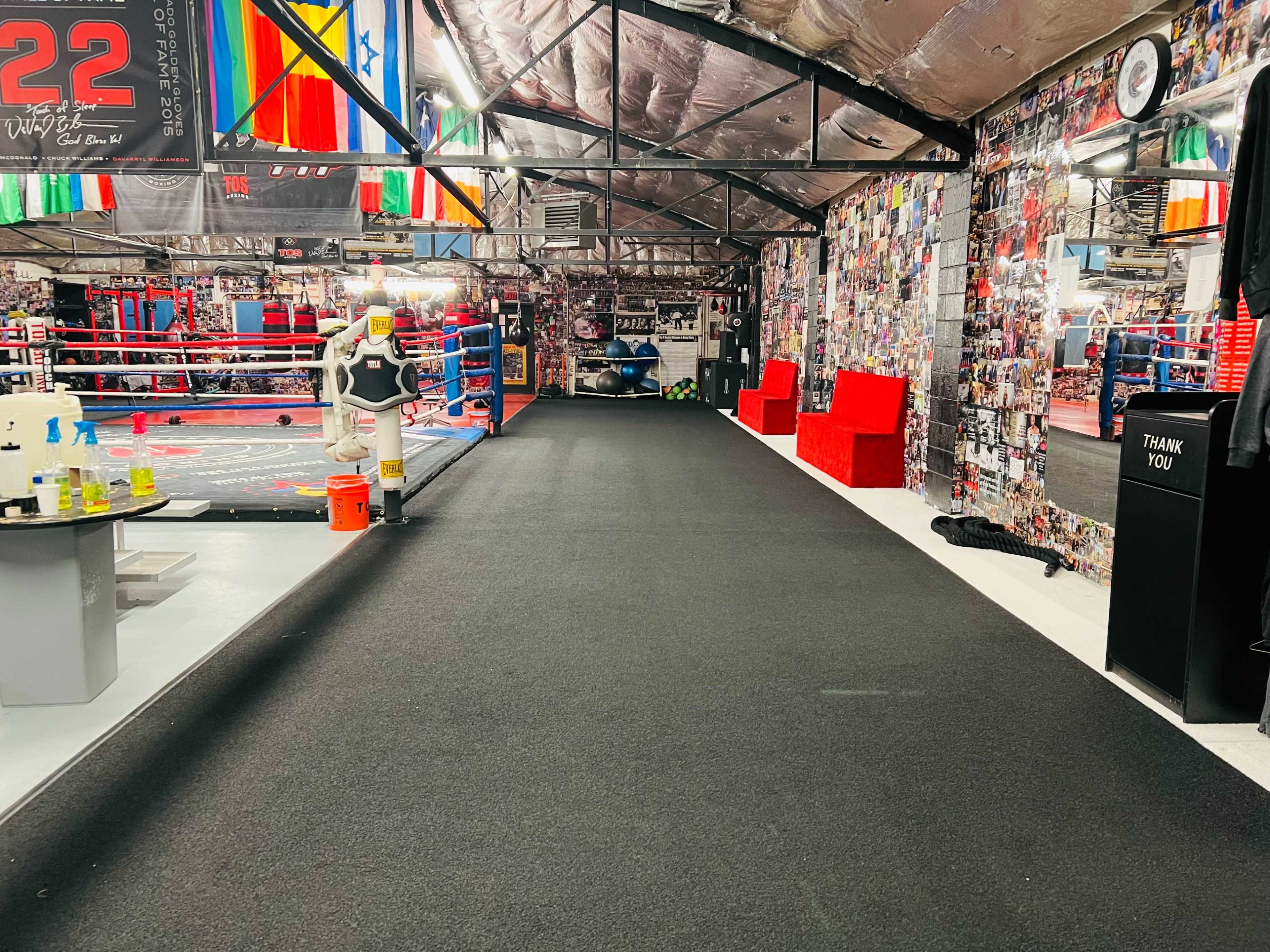 The image shows a spacious gym with a boxing ring, workout areas, and walls adorned with photographs and flags.