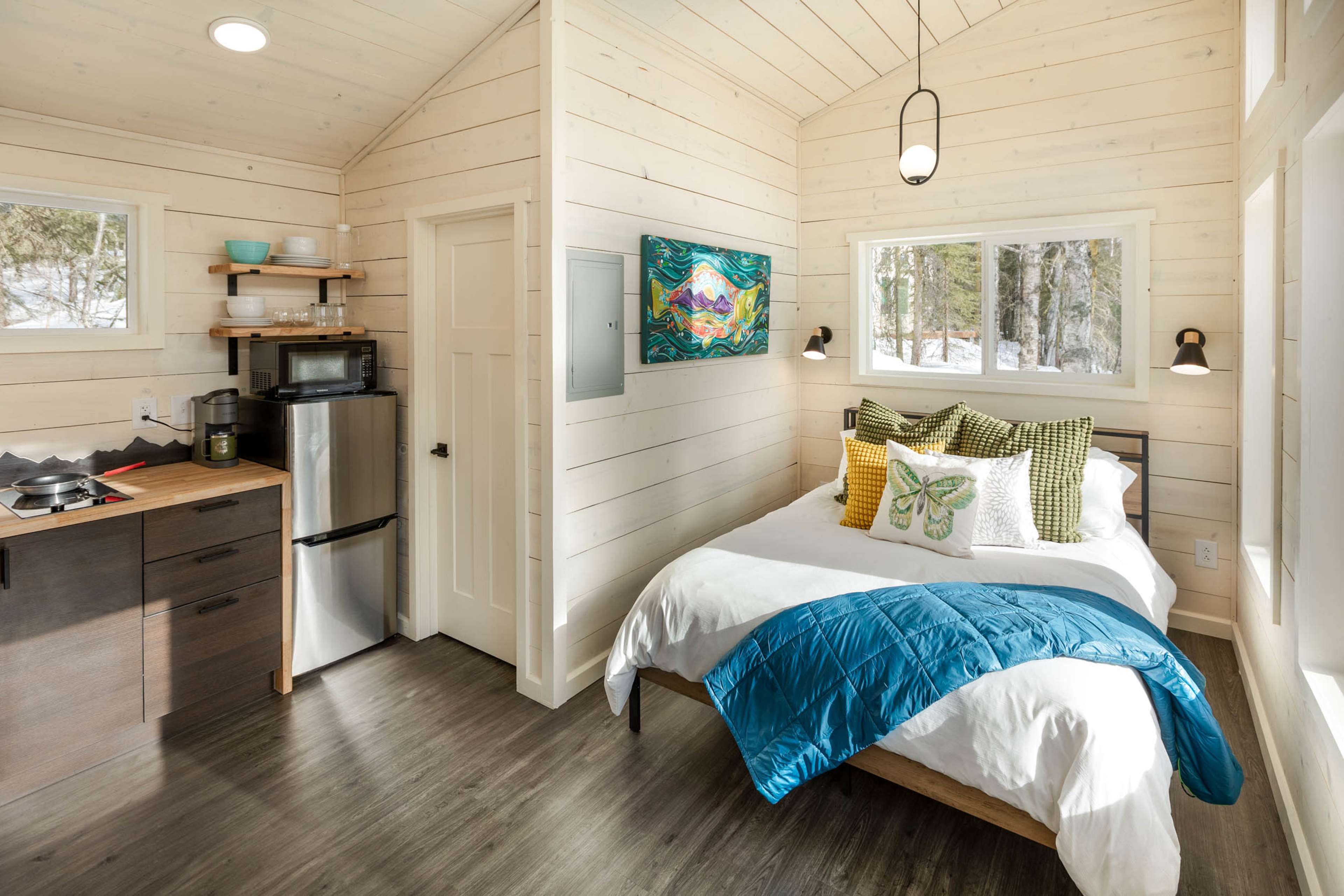 The image shows a small, modern cabin interior featuring a double bed with decorative pillows, a kitchenette with a refrigerator, and large windows letting in natural light.