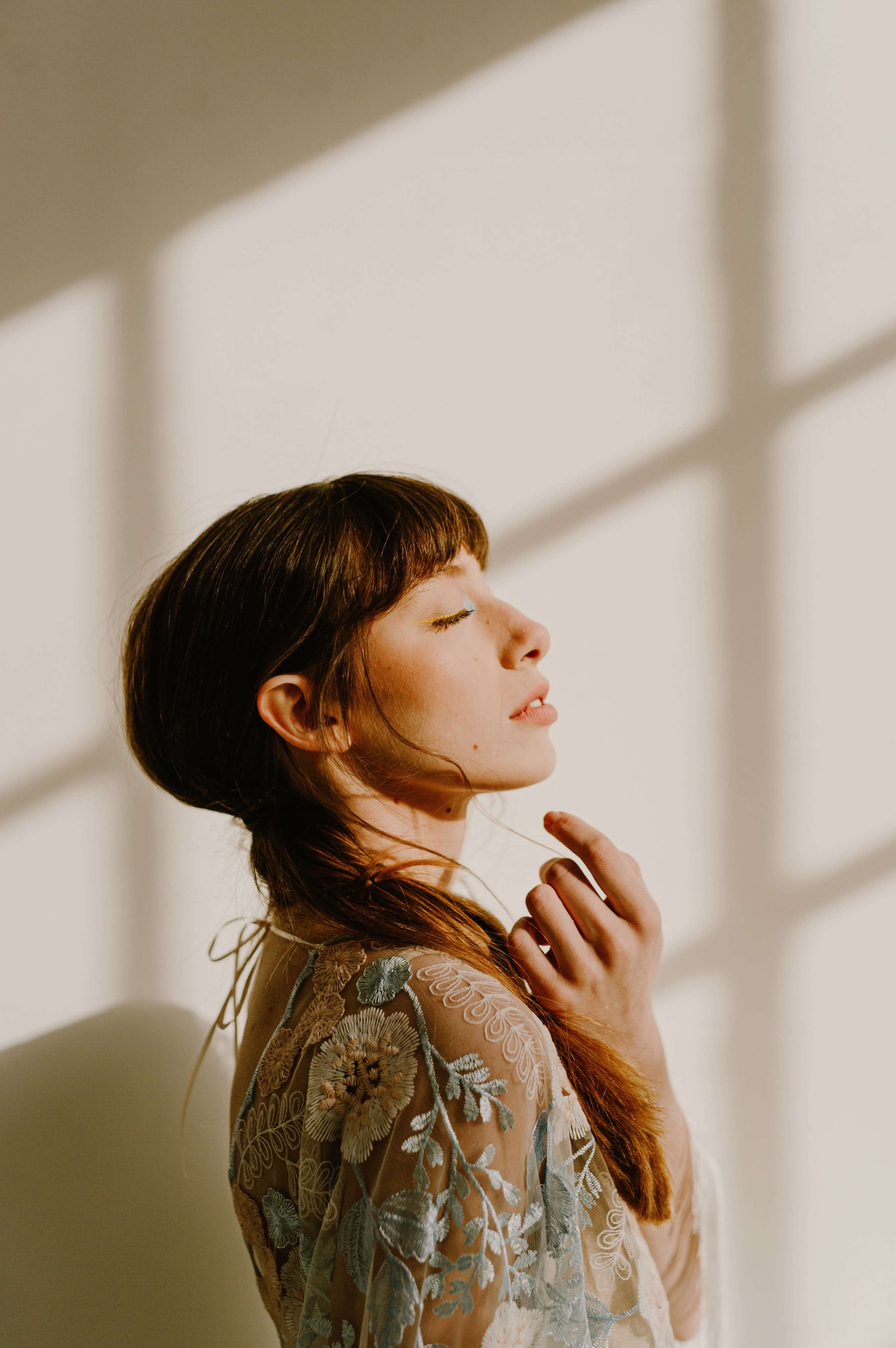 A woman with long hair stands in profile against a wall illuminated by sunlight, casting geometric shadows.