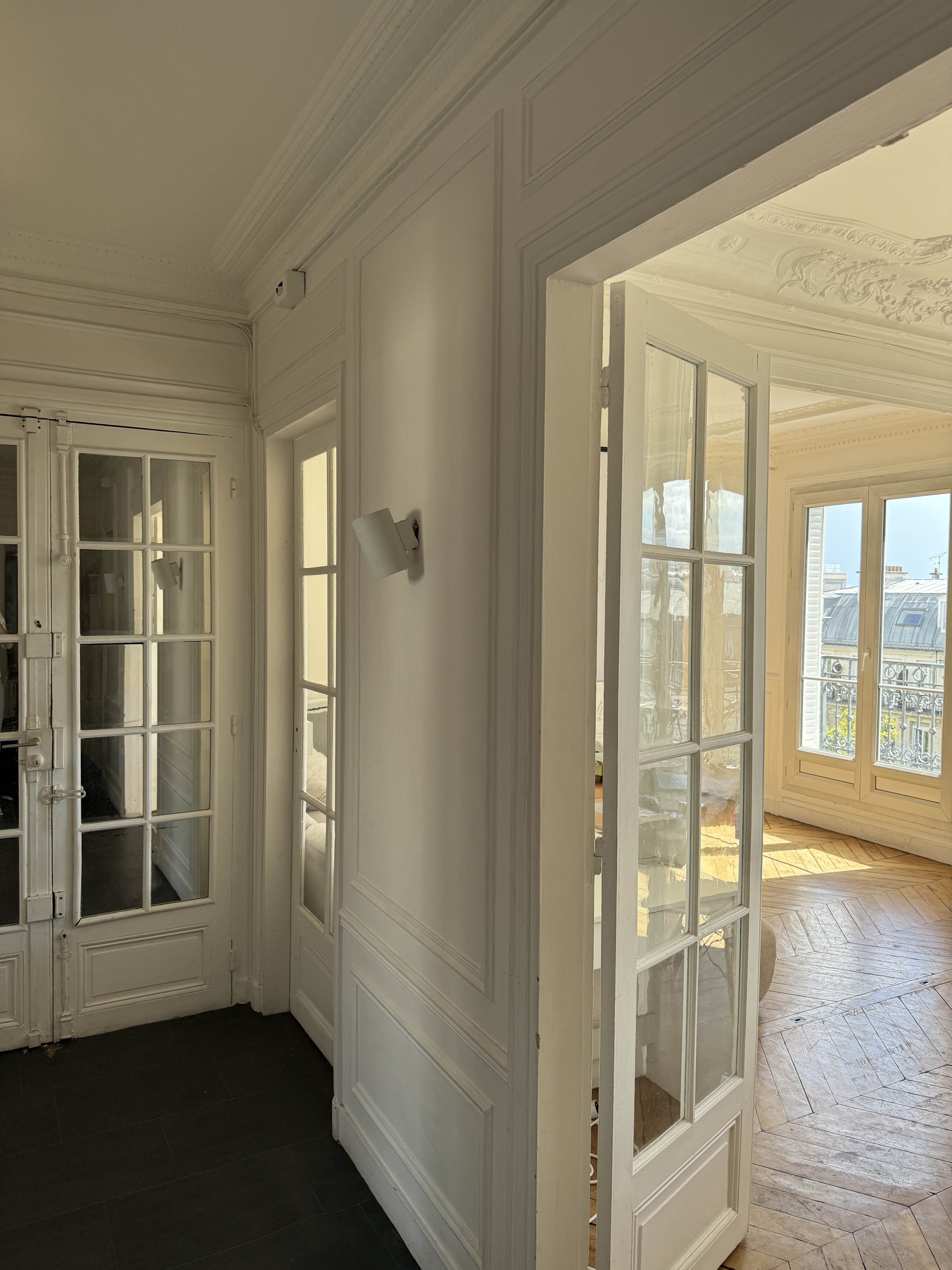 The image shows a sunlit hallway with white walls and French doors leading to a spacious room with large windows.