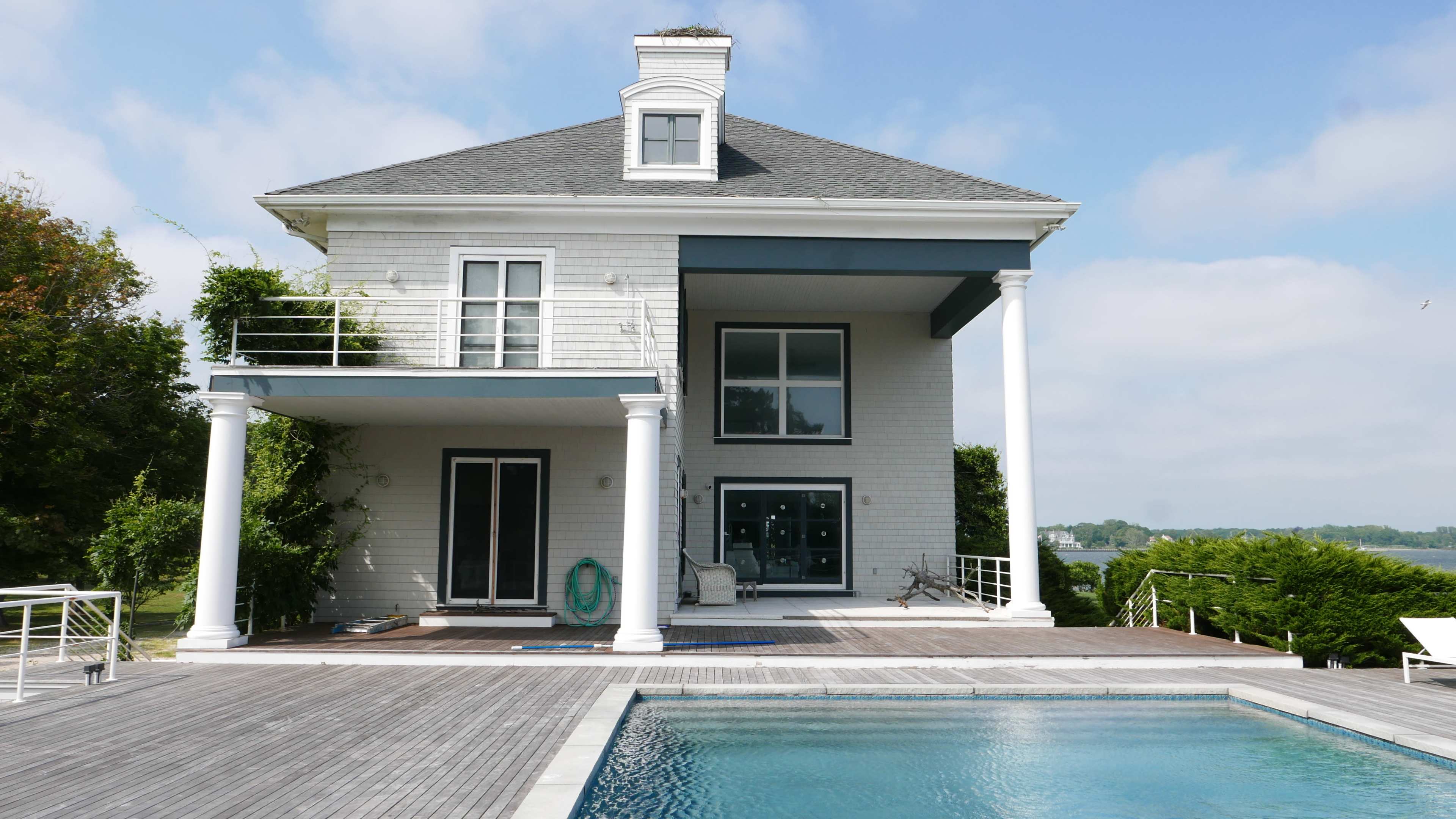 A two-story white house with large windows and a pool in the foreground is situated near a body of water.